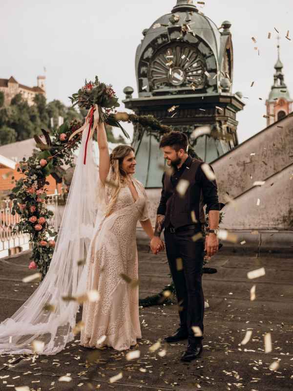 Bride and groom celebrating with confetti on a bridge with historic architecture in the background.