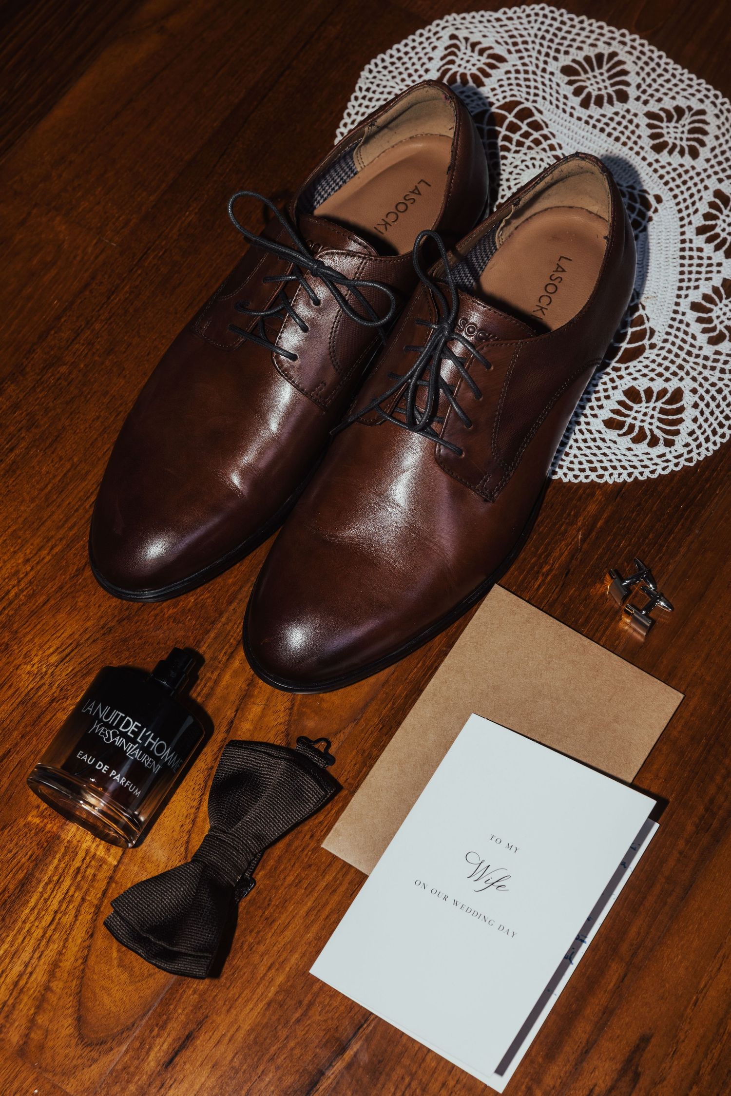 Brown leather dress shoes with bow tie, cologne, cufflinks, and wedding day card arranged on wooden floor.