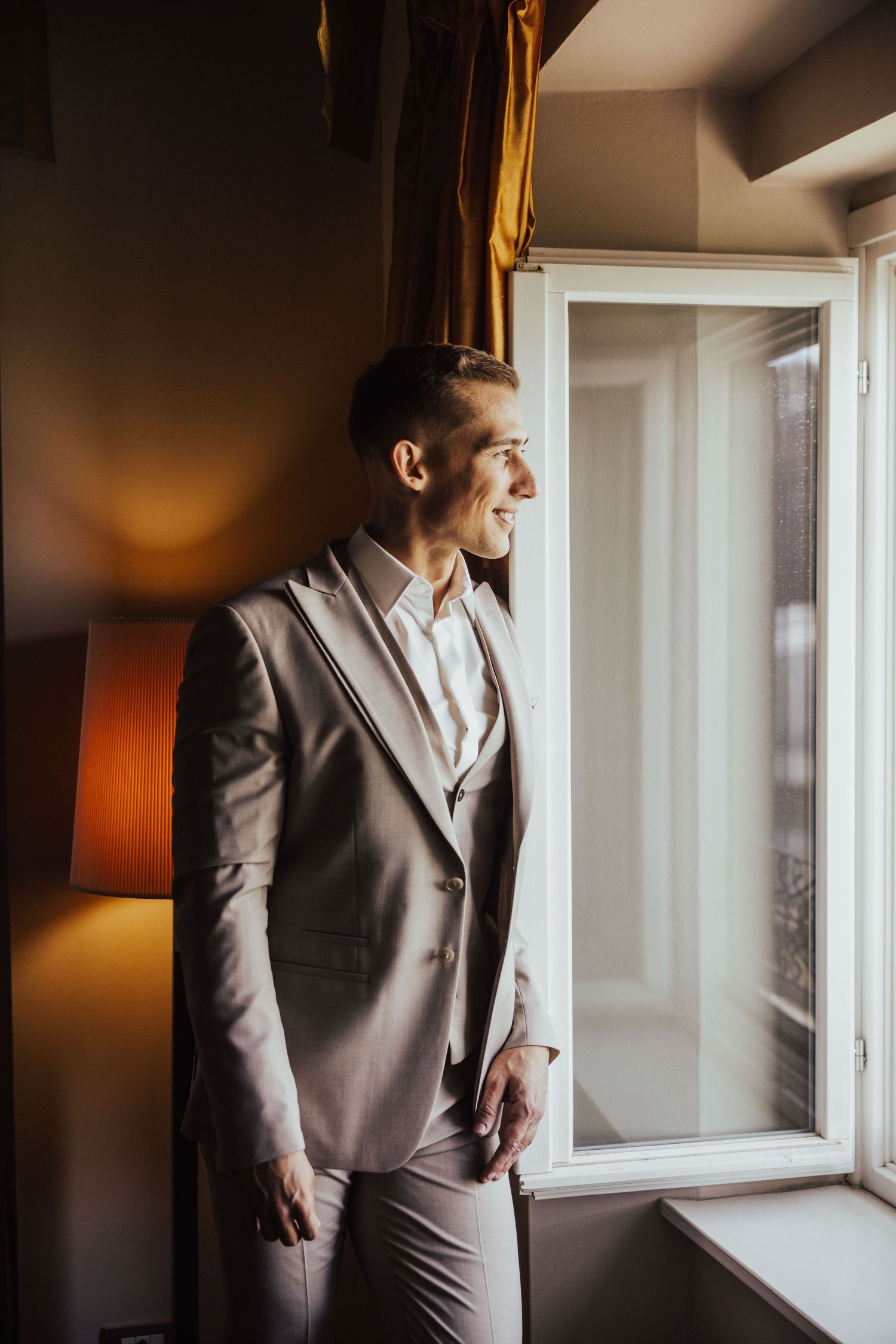 Smiling groom in beige suit standing by window in soft natural light on wedding day.
