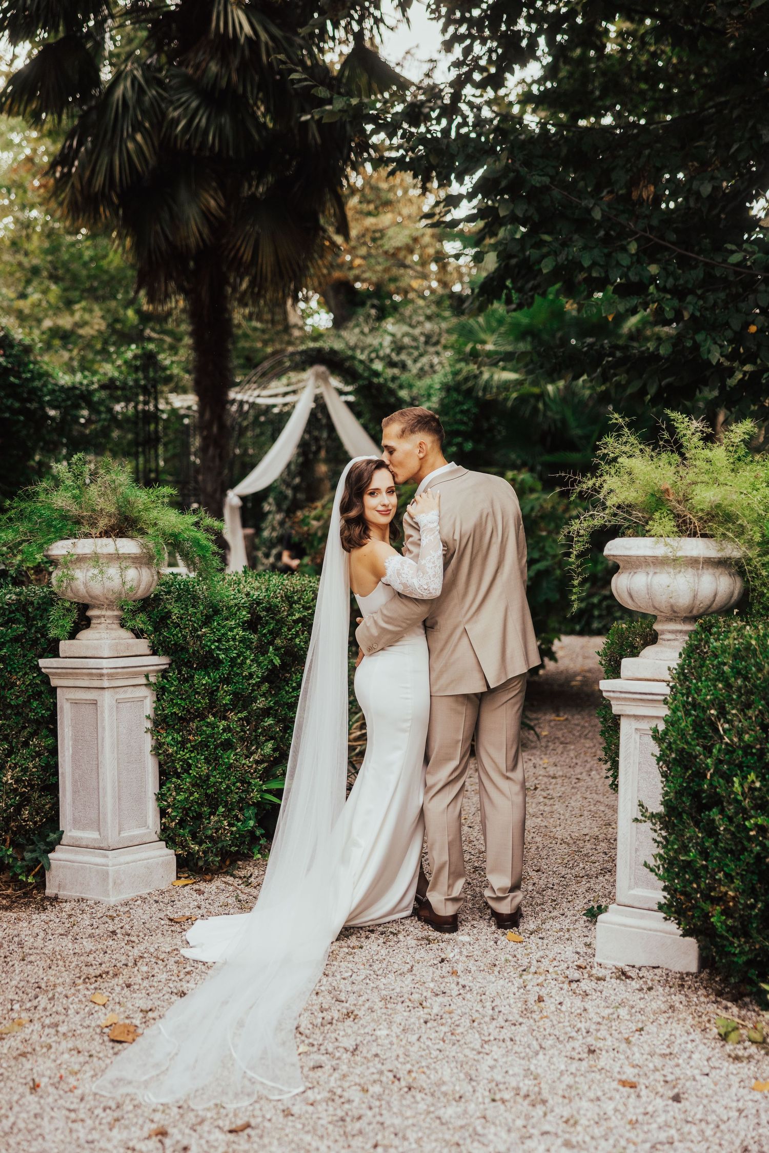 Bride and groom embrace in elegant garden with stone urns and lush greenery on wedding day.