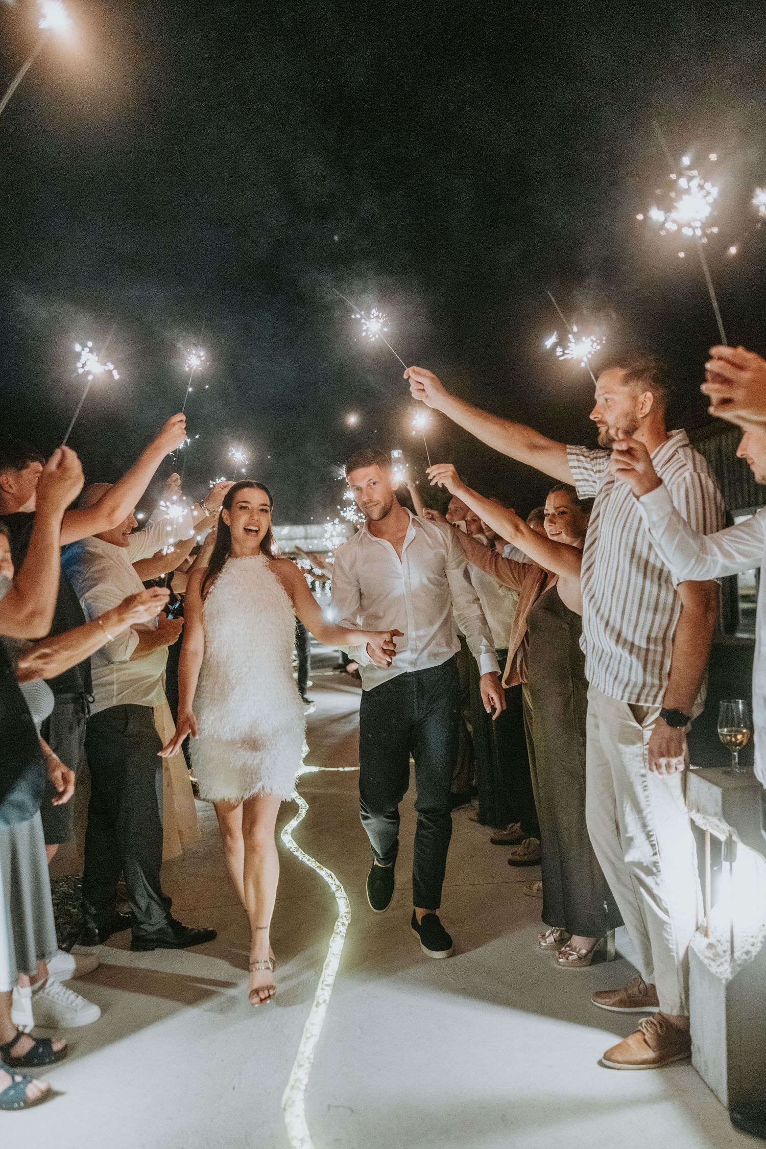 Happy bride and groom walk through glowing sparkler tunnel surrounded by celebrating wedding guests.