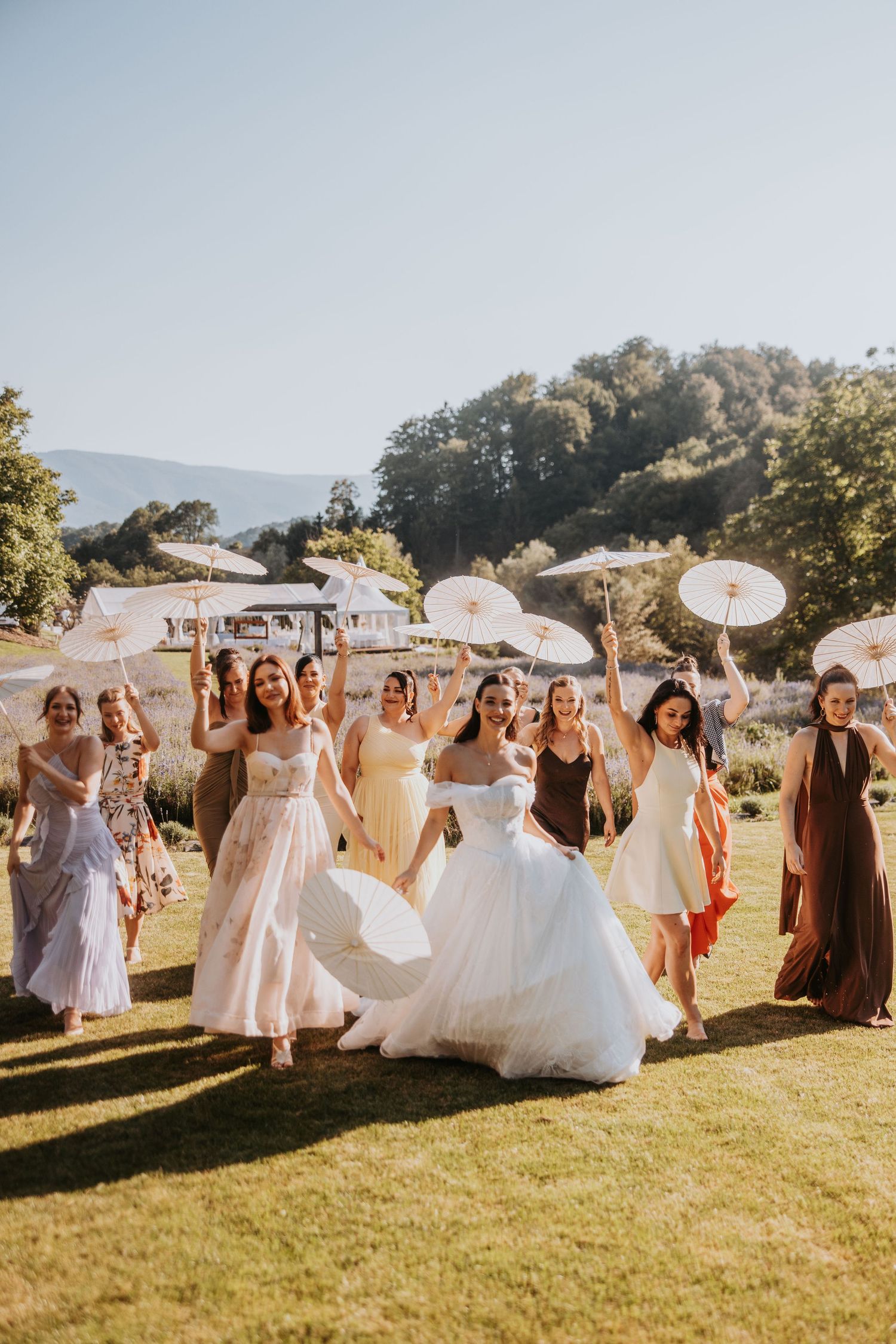 Bride in white gown walks with bridesmaids in colorful dresses holding parasols on sunny lawn.
