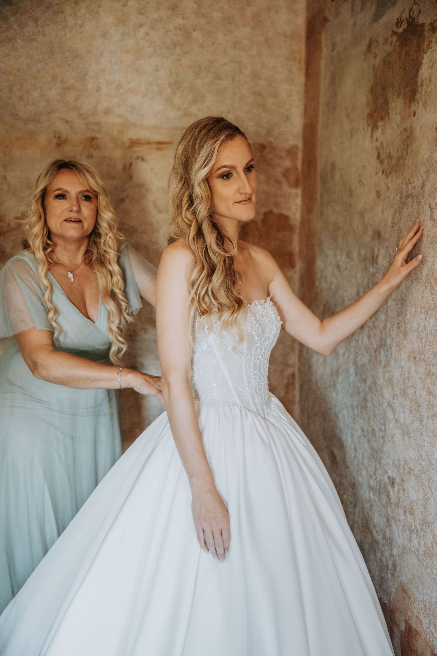 Mother helping bride into a strapless ballgown wedding dress against a rustic wall.
