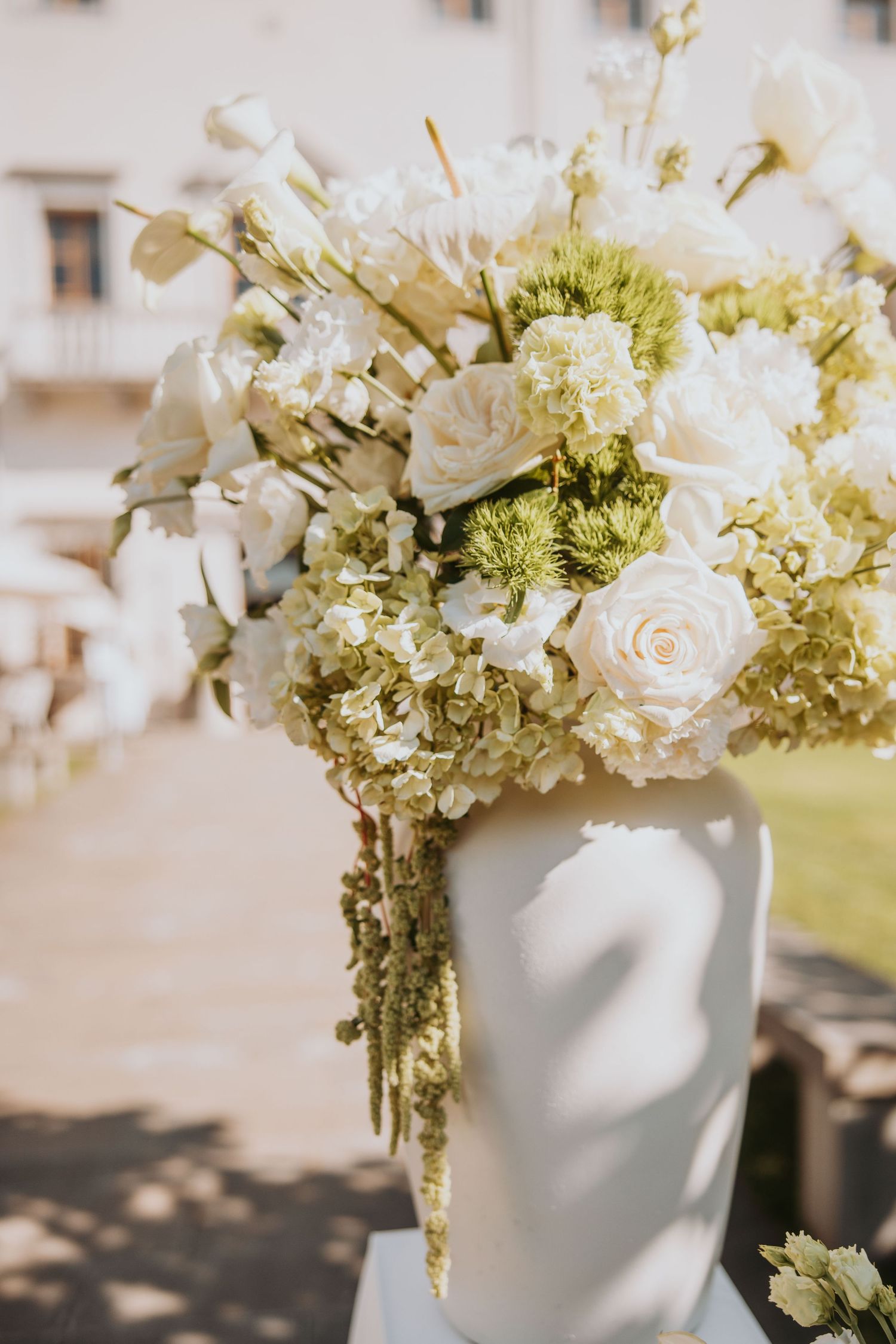 Lush white and green wedding bouquet with roses and hydrangeas in a white vase outdoors.