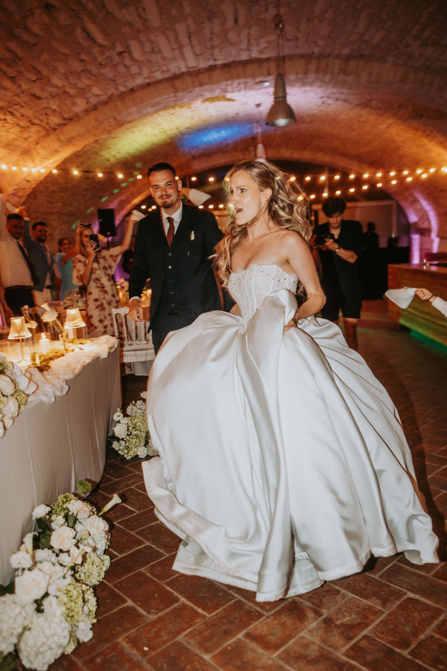 Bride in silver ballgown entering reception venue in rustic brick vaulted hall with string lights.