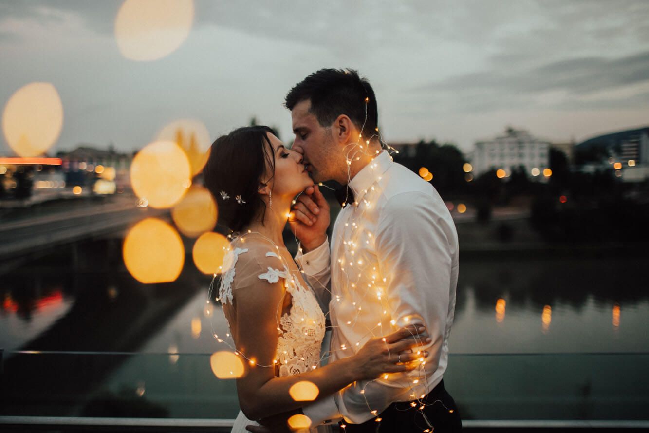 Newlyweds kissing wrapped in fairy lights with bokeh city reflections at dusk.