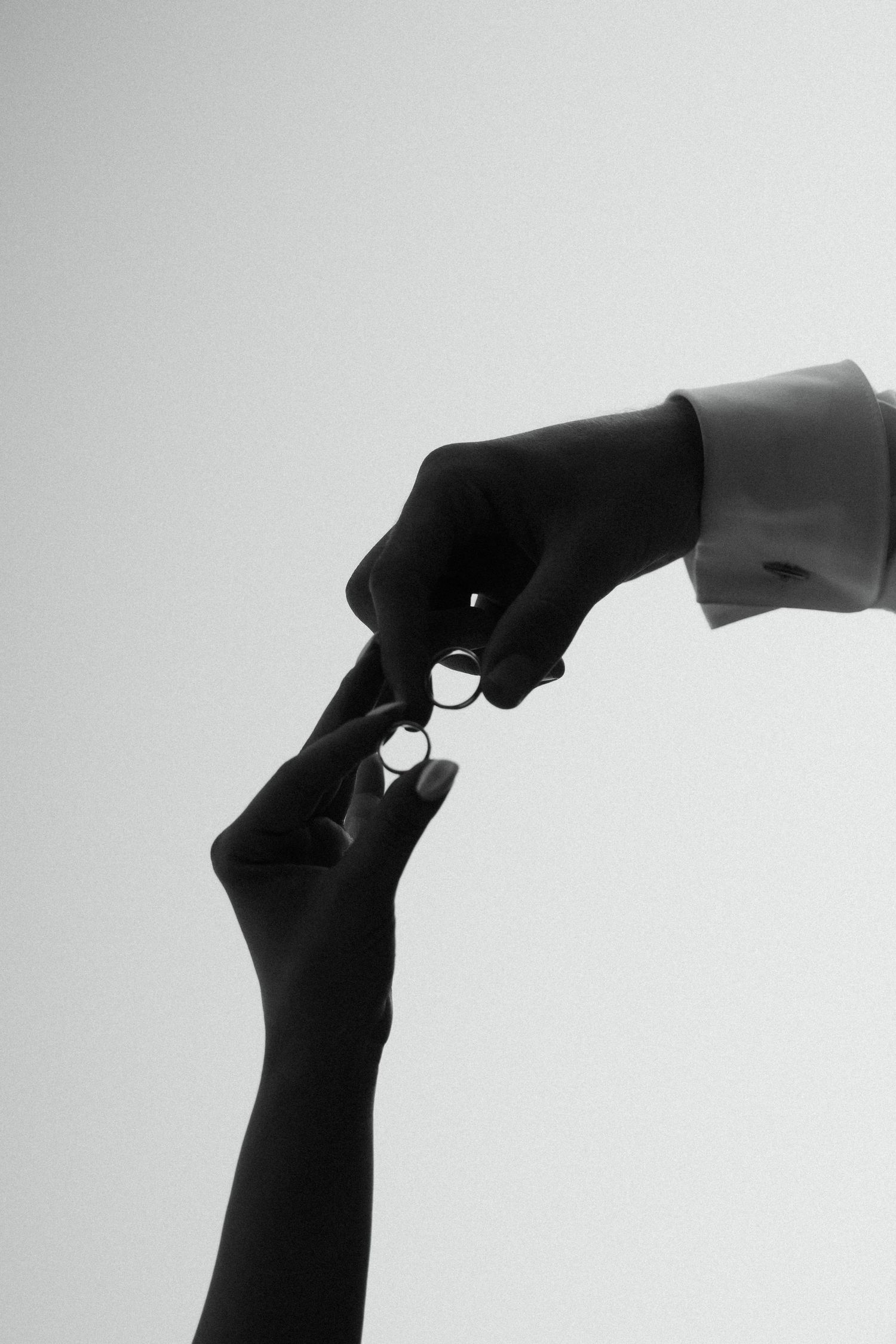 Black and white close-up of two hands exchanging wedding rings against a light background.