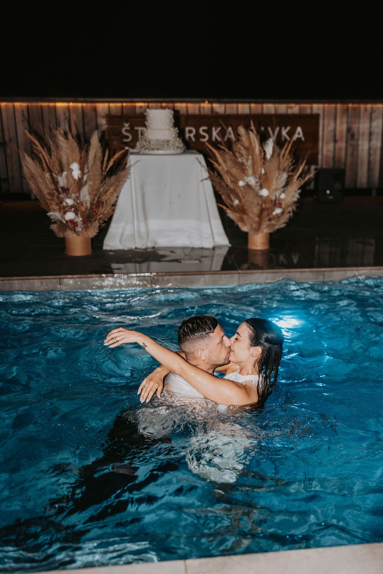 Couple kissing in illuminated pool at night with wedding cake display and dried floral arrangements in background.