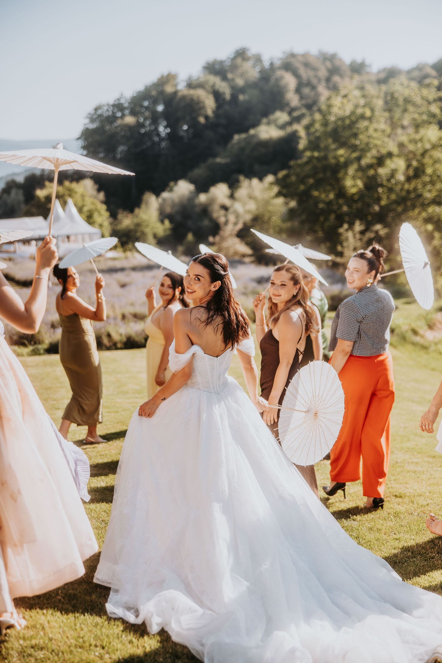 Bride in white ball gown dancing outdoors with guests holding parasols on sunny day.