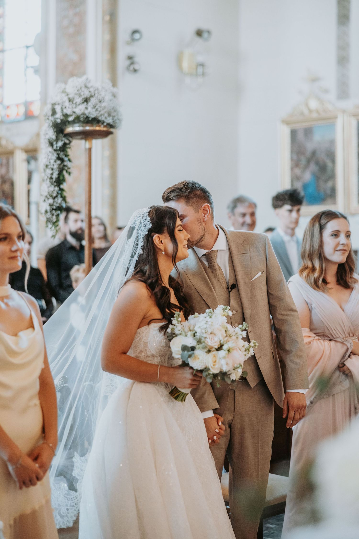 Bride and groom share a kiss at the altar during their church wedding ceremony, surrounded by guests and floral decorations.