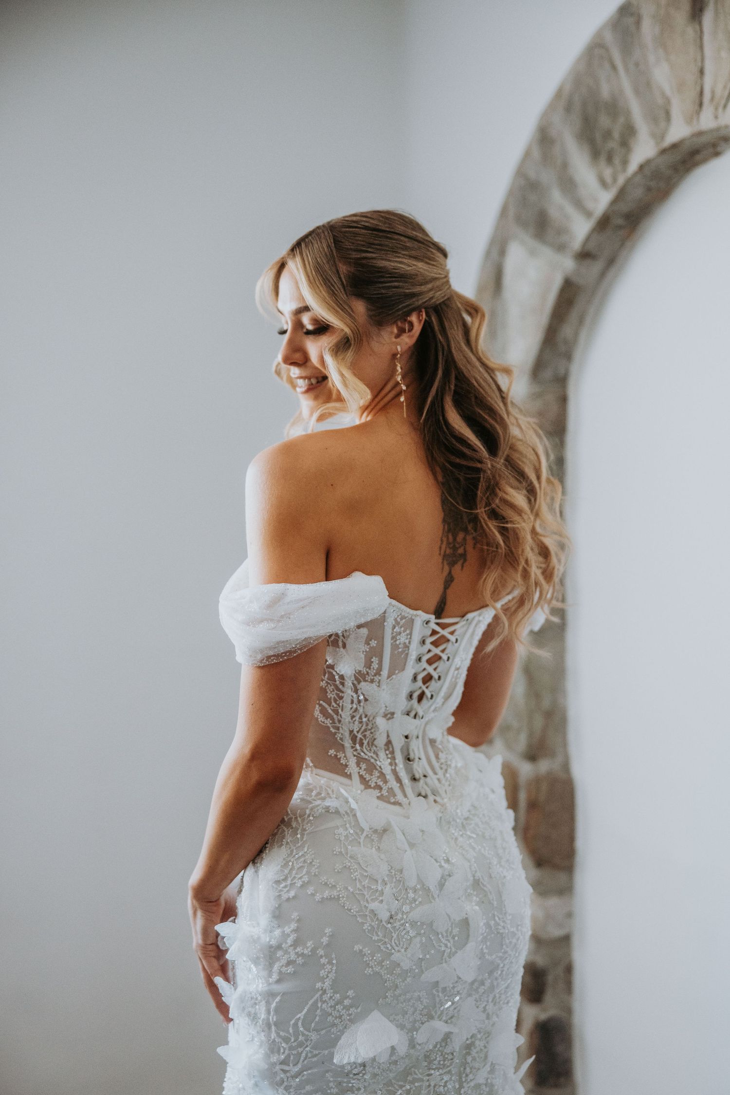 Bride in elegant off-shoulder lace wedding gown with corset back smiling near stone arch.