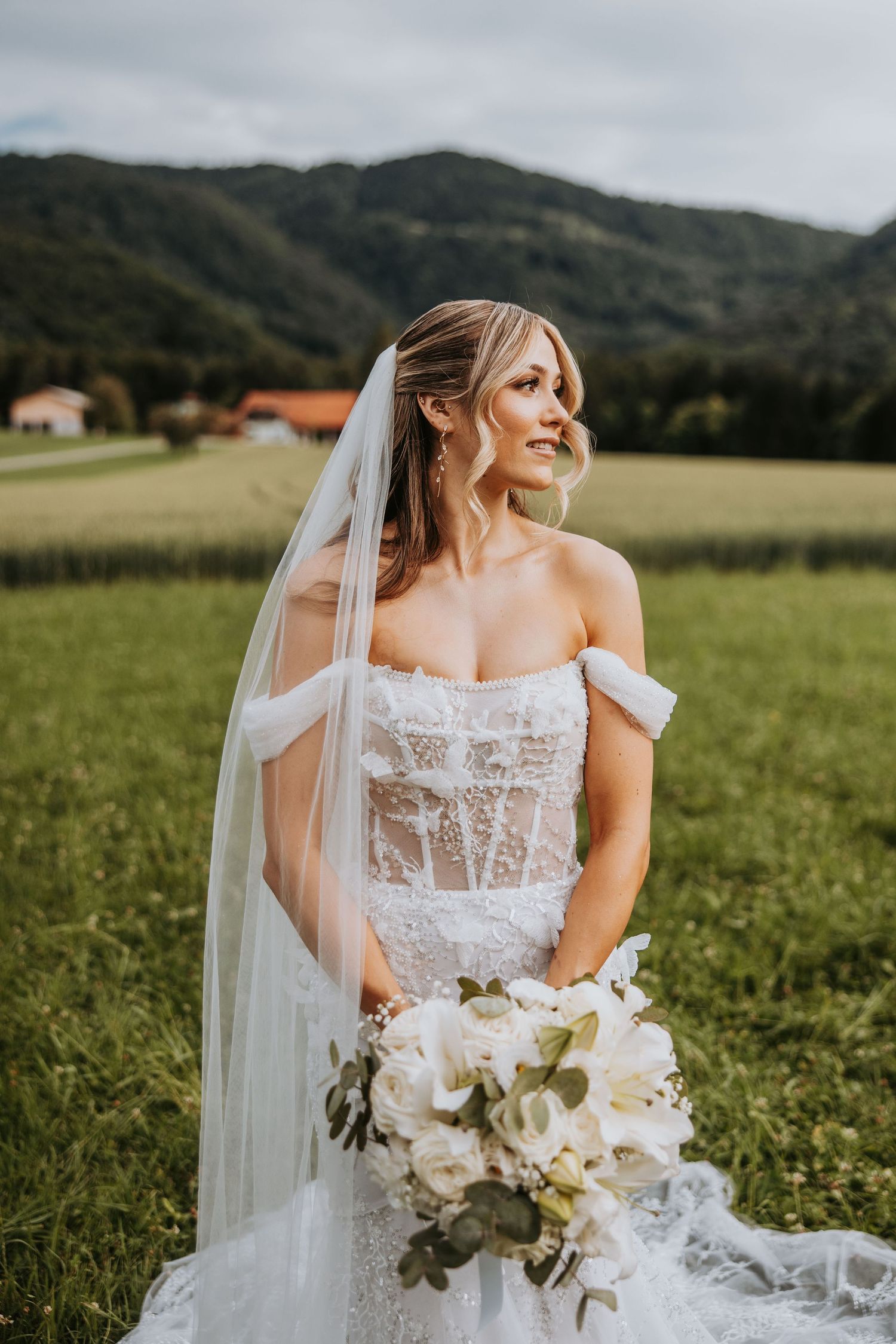 Bride in lace off-shoulder gown holding white bouquet in a green meadow with mountain backdrop.