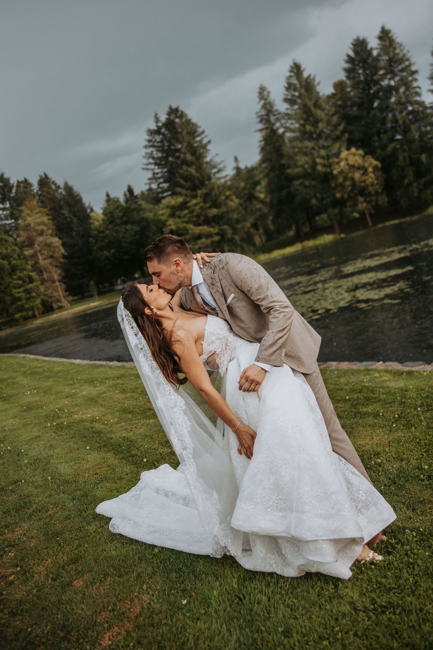 Groom dipping bride for passionate kiss on green lawn with dramatic stormy sky backdrop.