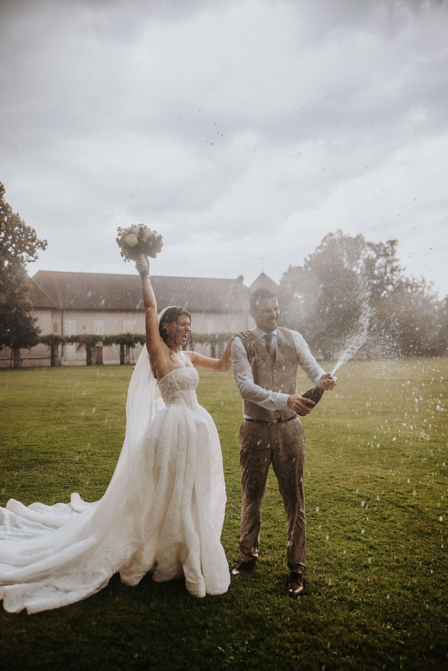 Joyful bride raises bouquet as groom pops champagne bottle on green lawn during outdoor wedding celebration.