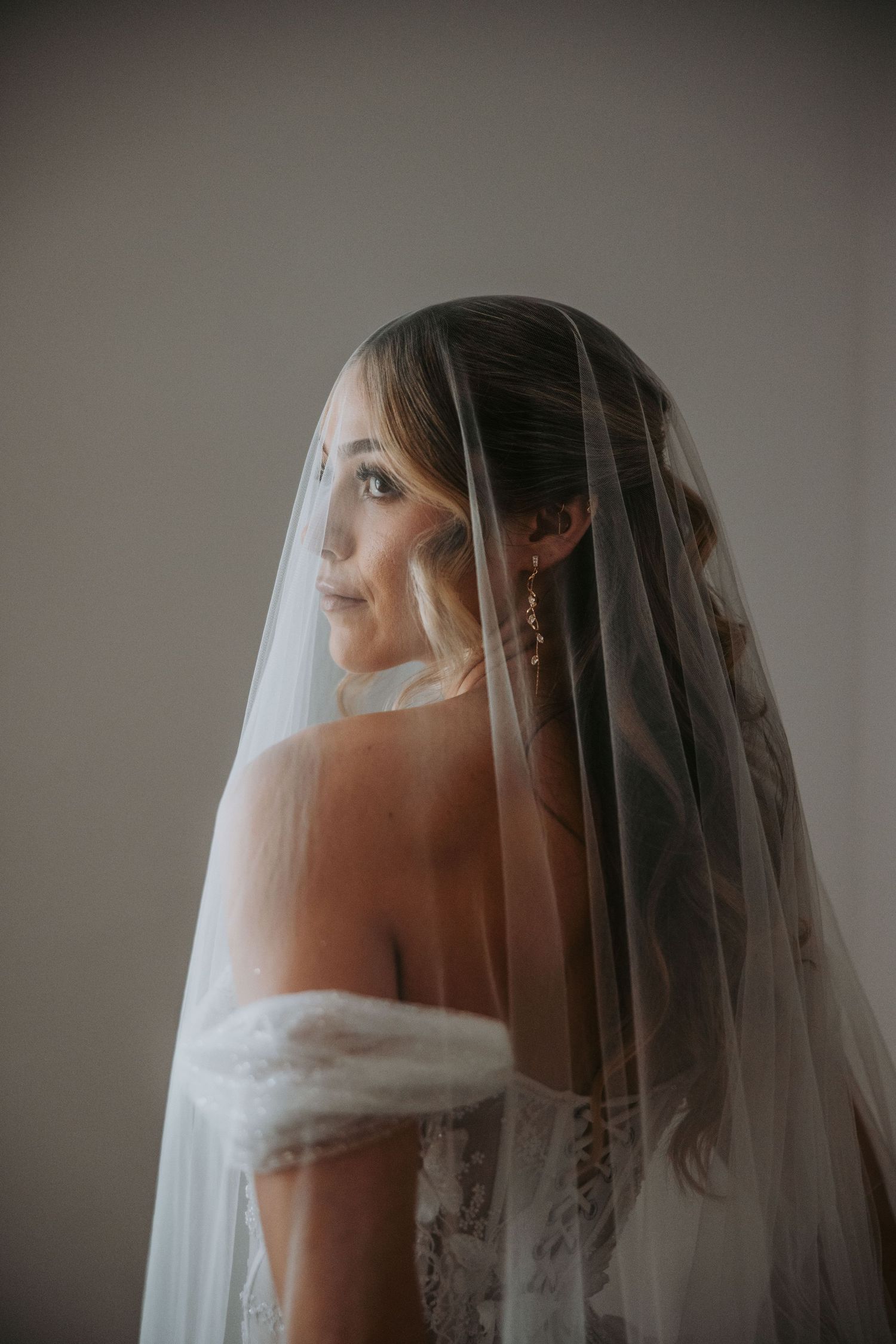 Elegant bride in off-shoulder gown gazing sideways under a sheer veil in soft studio lighting.