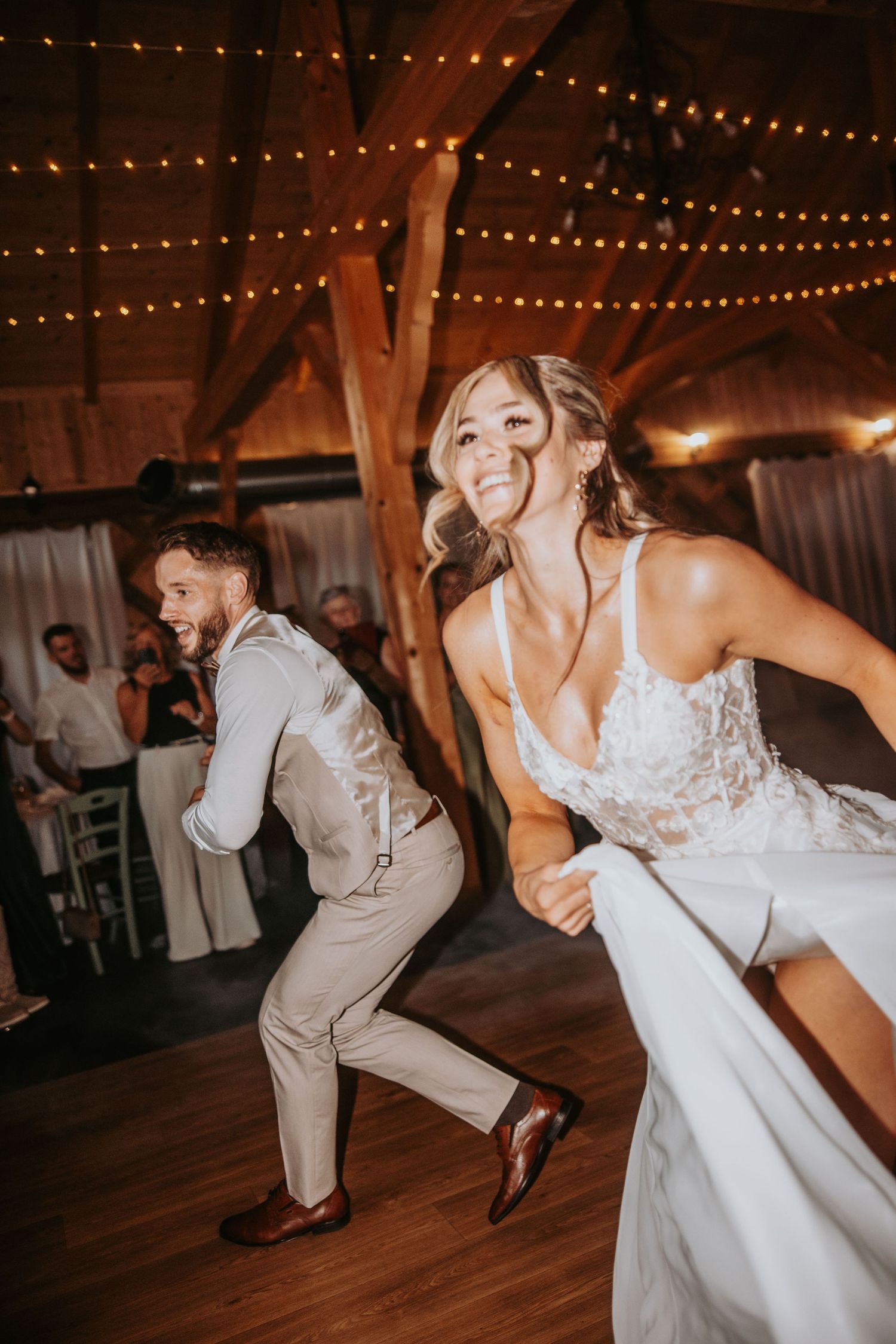 Joyful bride and groom dance energetically in a warmly lit rustic barn wedding reception venue.