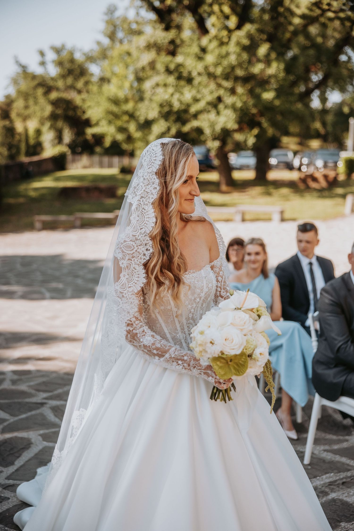 Radiant bride in lace wedding gown and veil holds white bouquet during outdoor ceremony on sunny day.