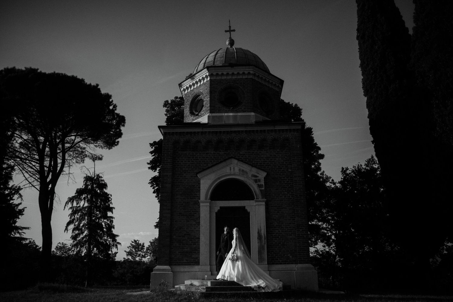 Dramatic black and white photo of a bride in a white gown standing before a historic domed chapel.