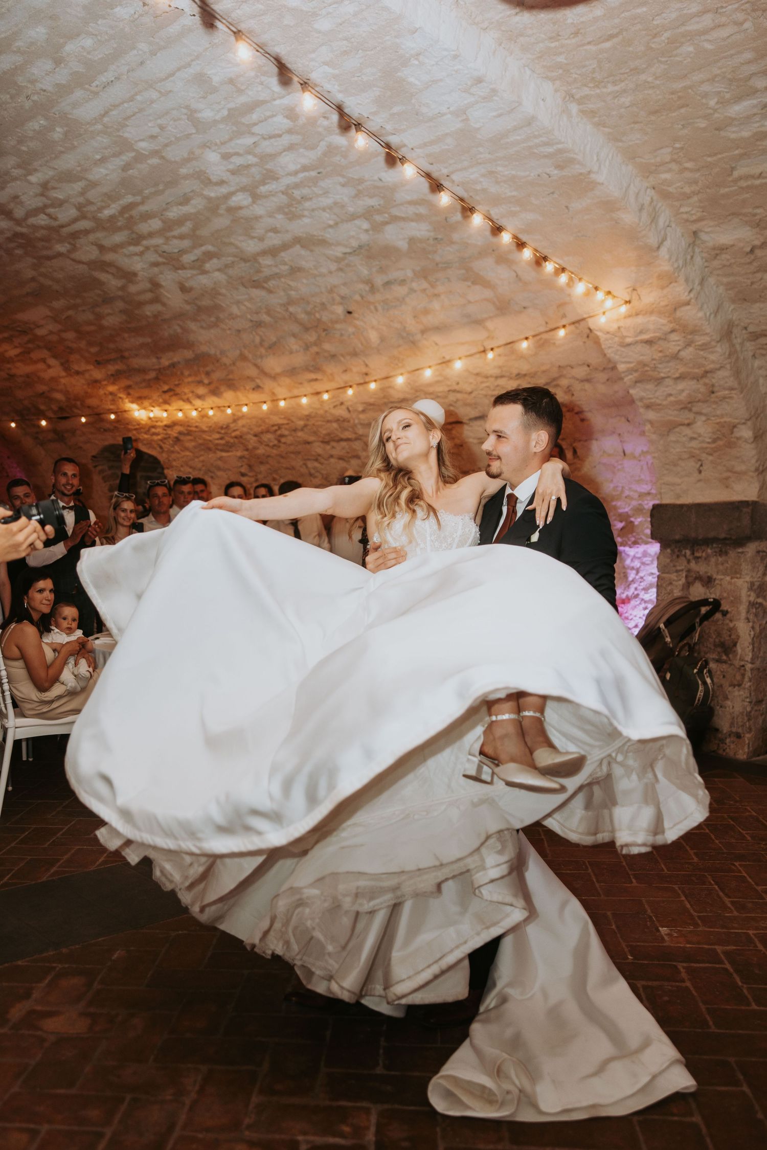 Groom lifting bride during first dance in rustic stone venue with string lights.