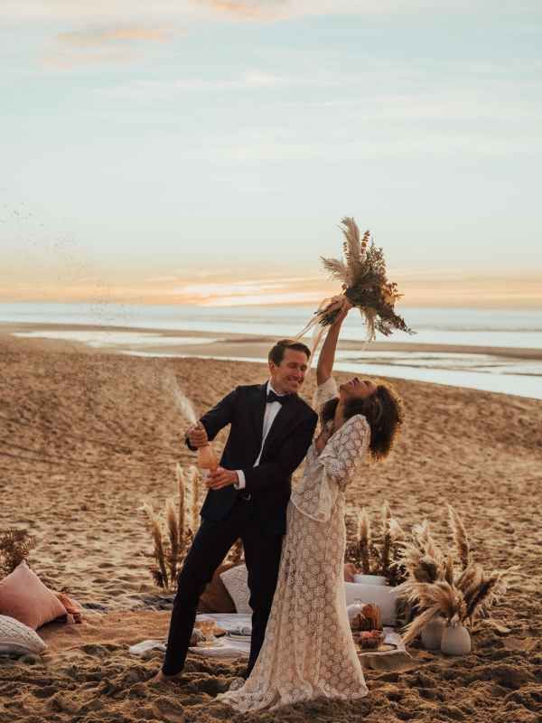 A joyful couple celebrates their beach wedding at sunset, with the groom popping champagne and the bride raising her bouquet.