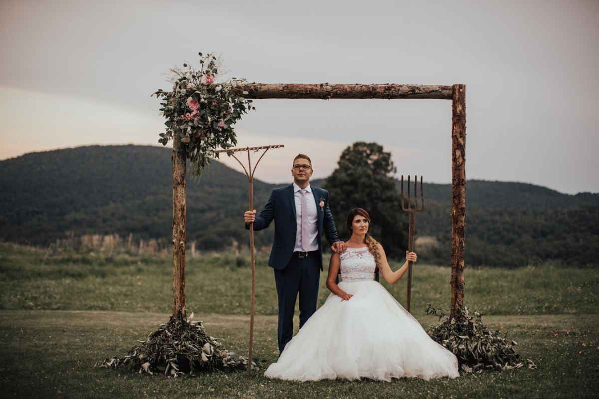 Bride and groom posing under rustic wooden arch in scenic countryside wedding ceremony.
