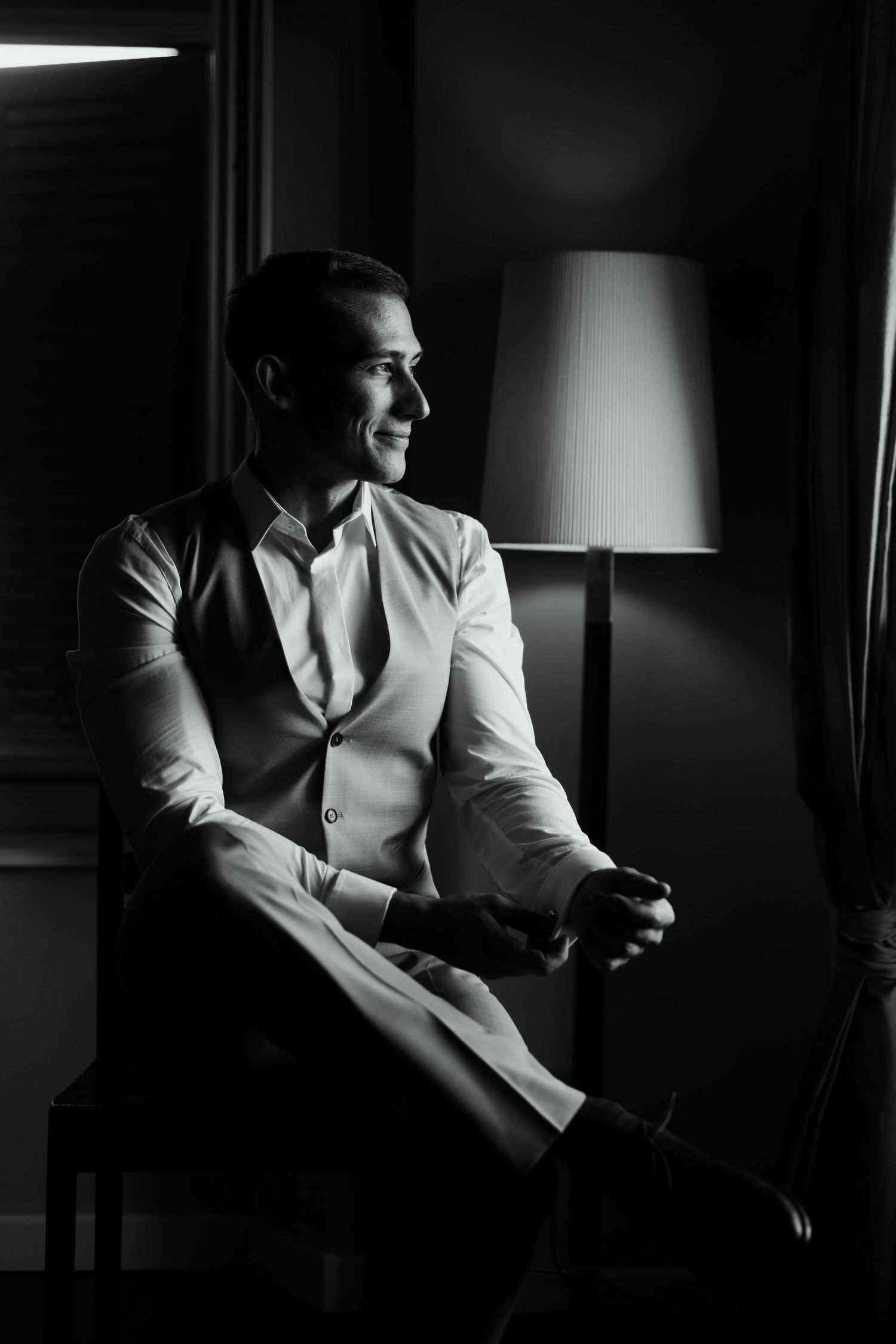 Stylish groom in white vest and shirt seated by a lamp in dramatic black and white portrait photo.