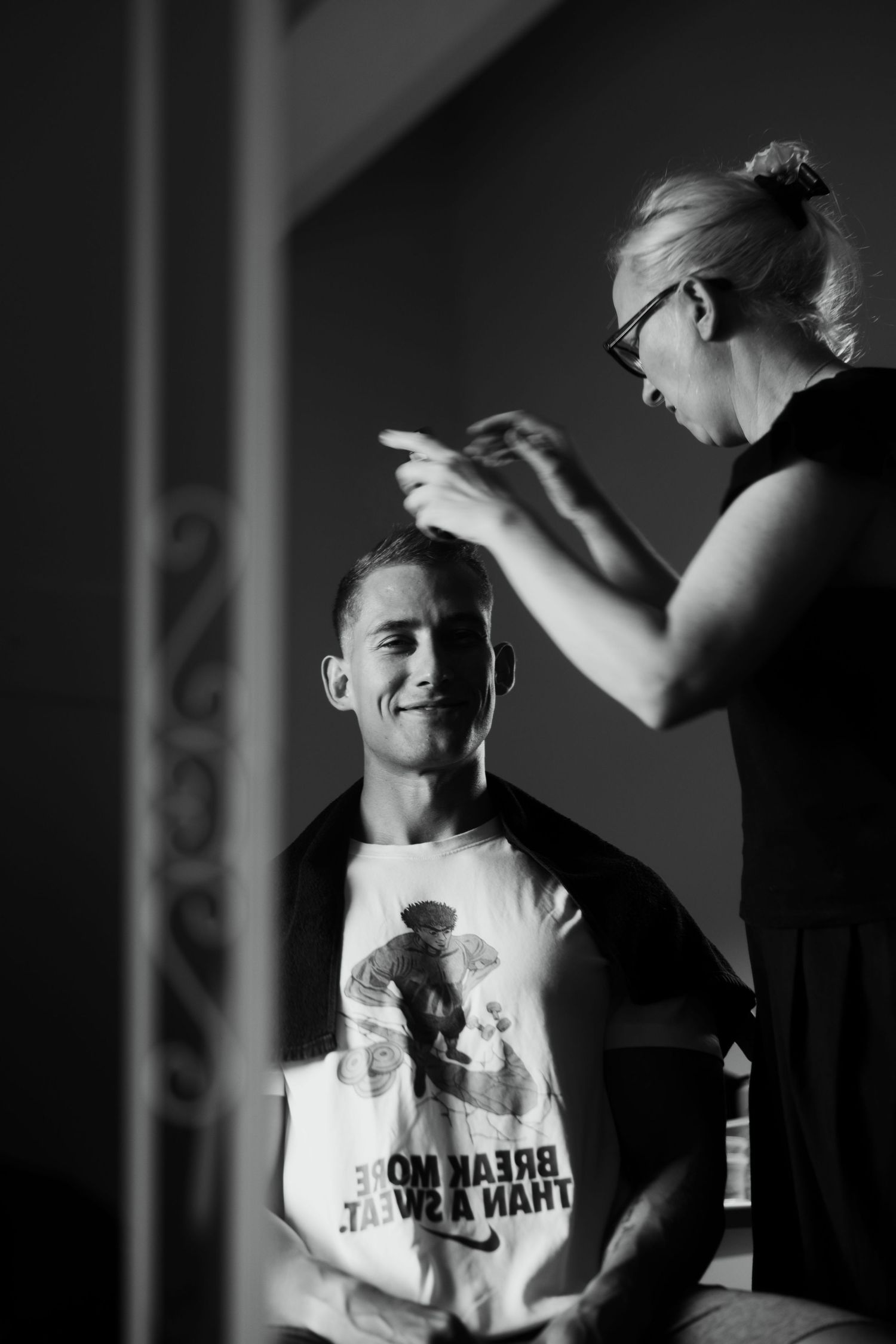 Smiling groom gets hair styled by blonde hairstylist during wedding preparations in black and white photo.