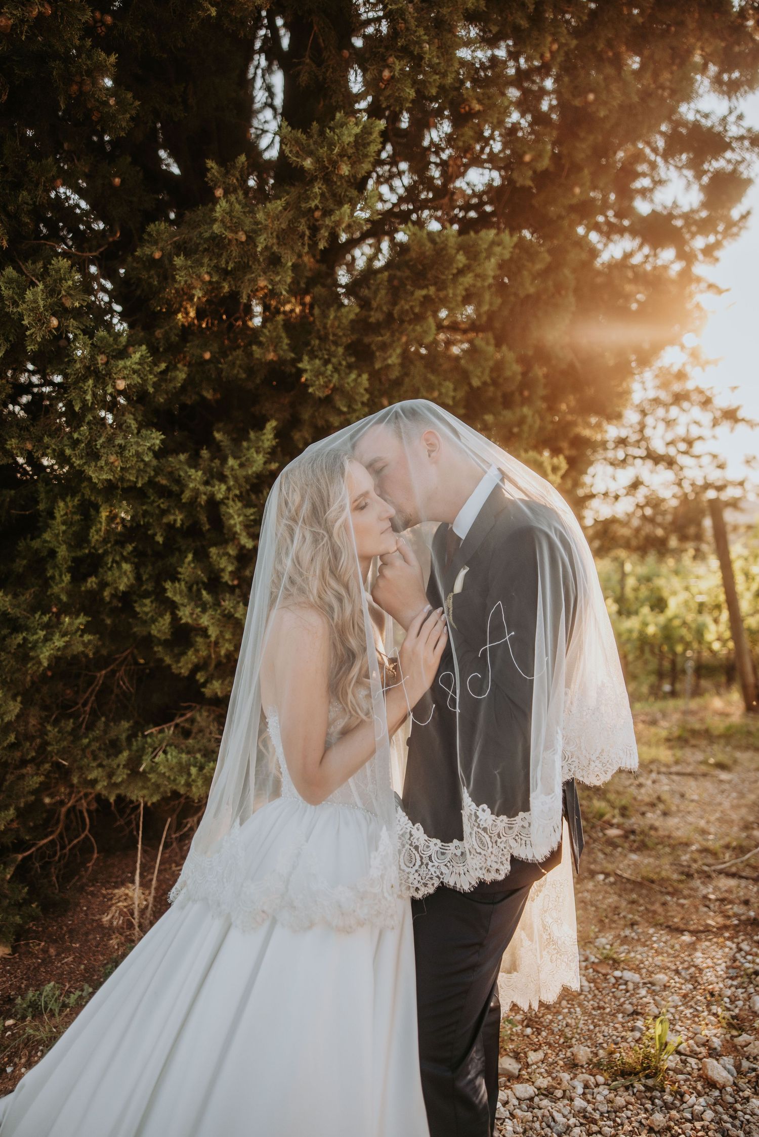 Bride and groom sharing a romantic kiss under lace monogrammed veil with golden sunset light filtering through trees.
