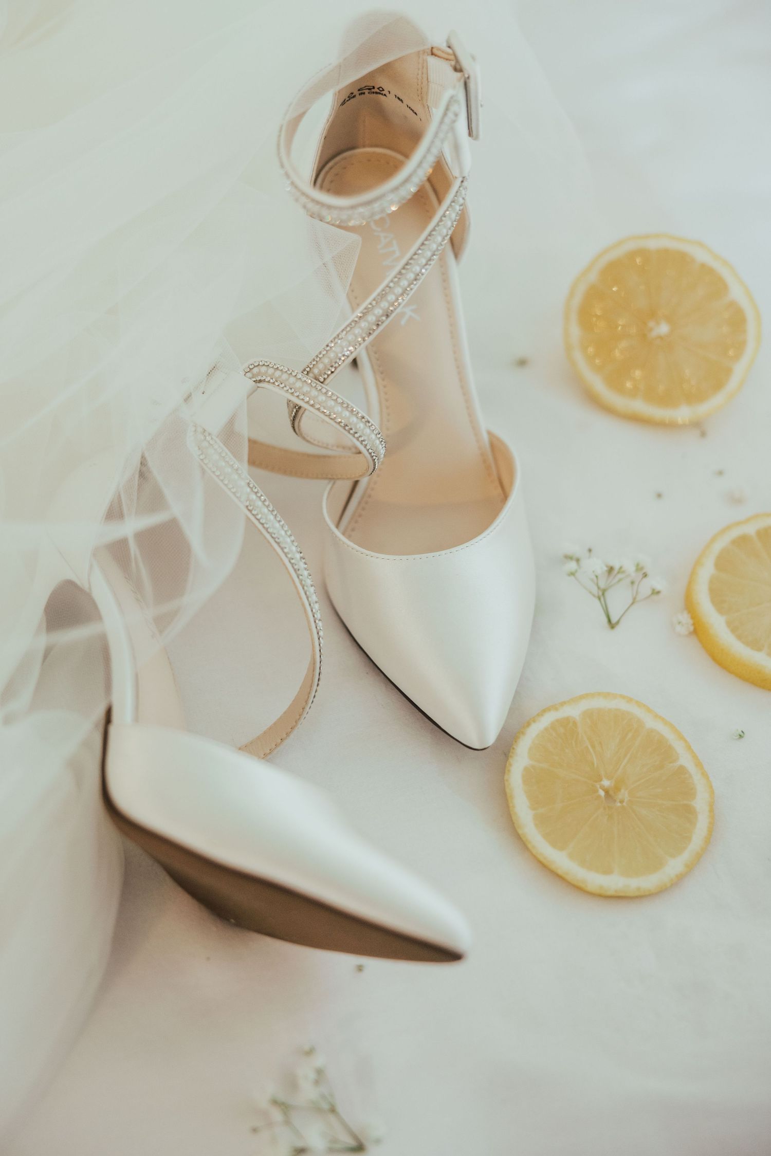 Elegant white bridal heels with crystal ankle straps displayed beside fresh lemon slices on a white surface.