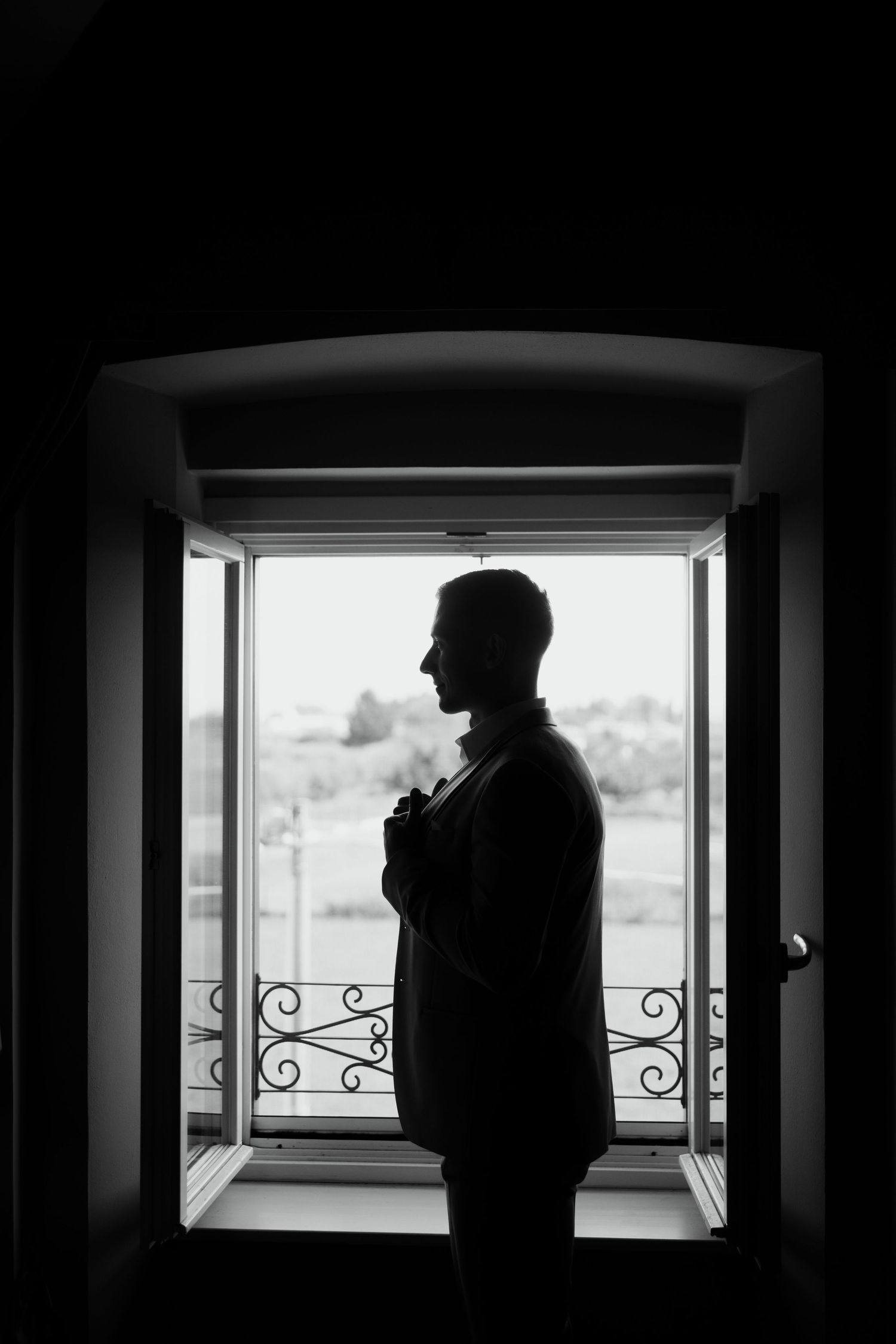 Silhouette of groom standing at open window in black and white wedding portrait photography.