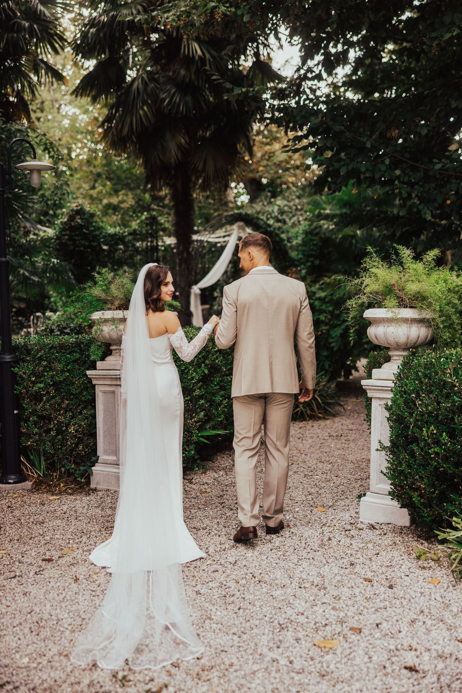 Bride and groom walking hand in hand through elegant garden pathway with stone urns and tall trees.