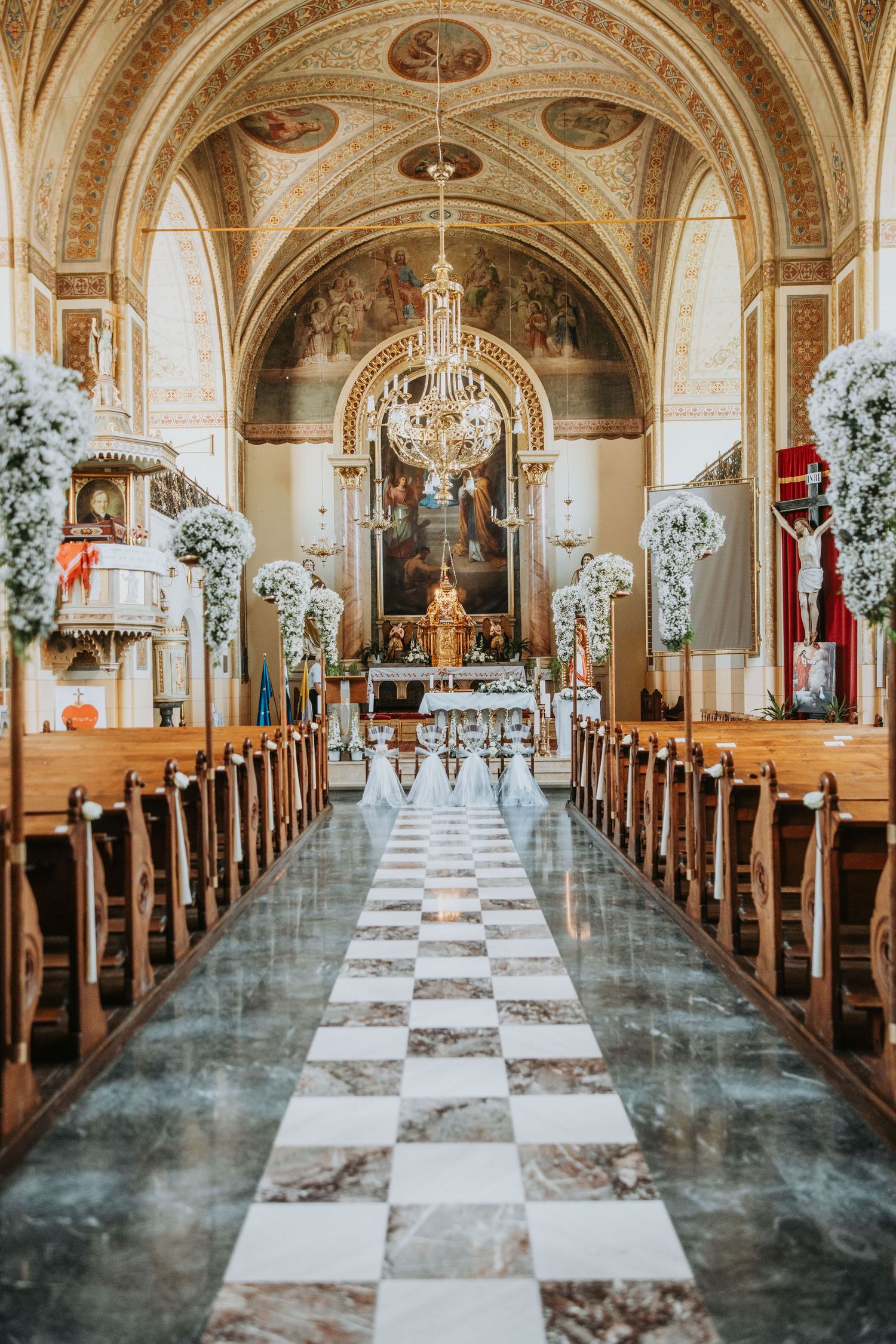 Elegant church interior with checkered aisle, wooden pews, white floral arrangements, ornate frescoed ceiling, and golden altar.
