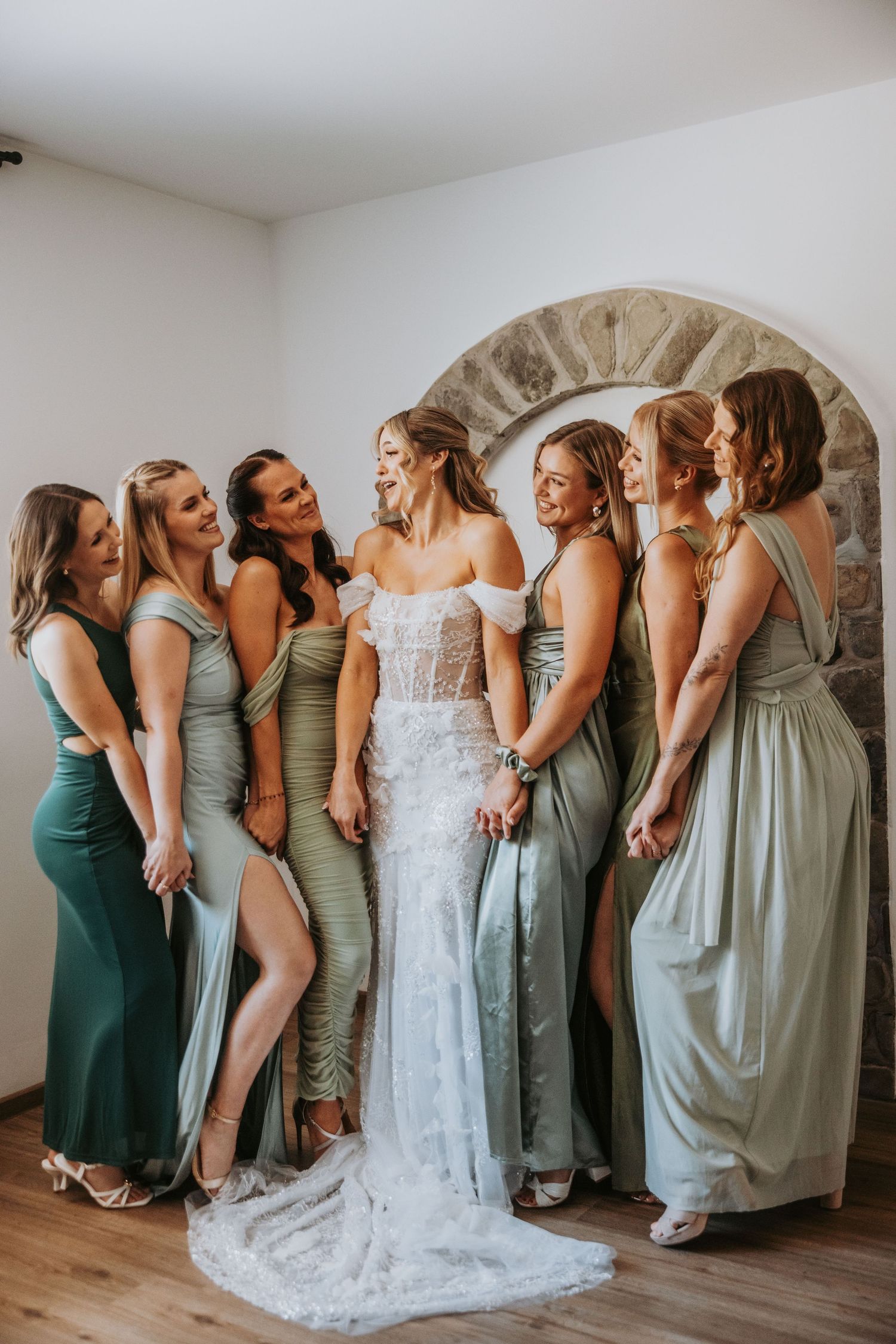 Bride in white lace gown surrounded by bridesmaids in sage green dresses posing together indoors.