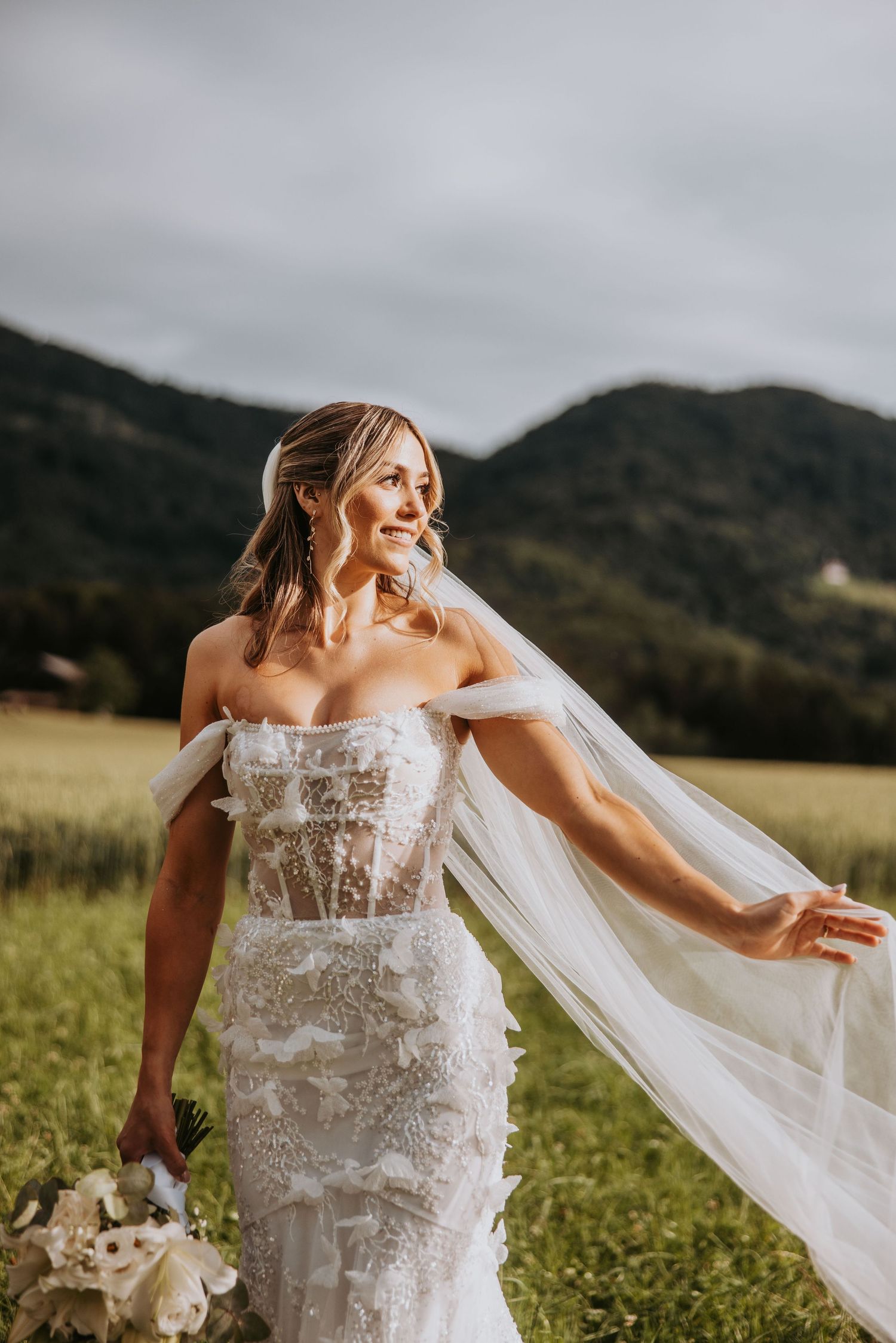 Smiling bride in lace off-shoulder gown holding veil open in a sunlit field with mountain scenery.