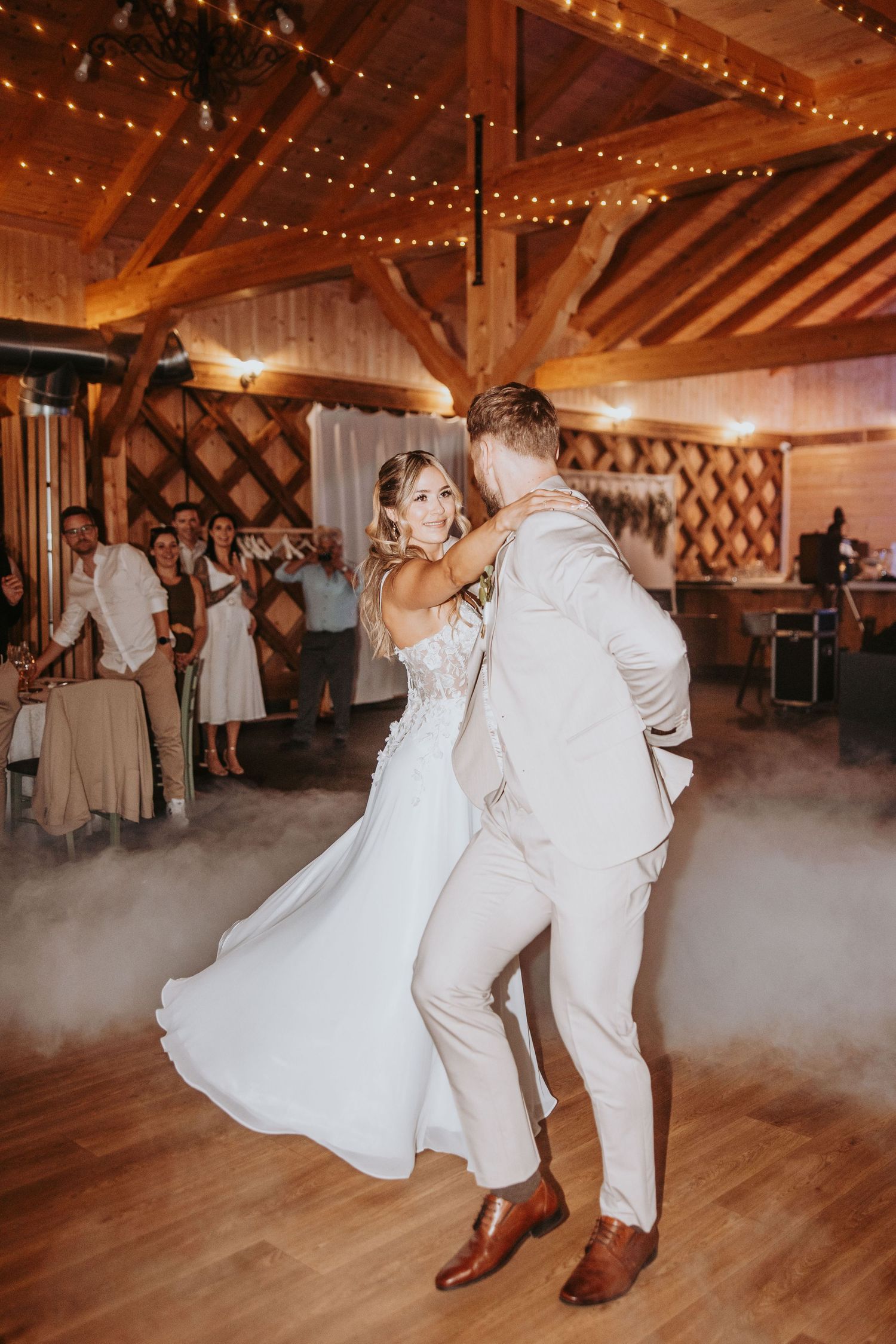 Bride and groom share first dance in rustic wooden venue with string lights and fog effect.