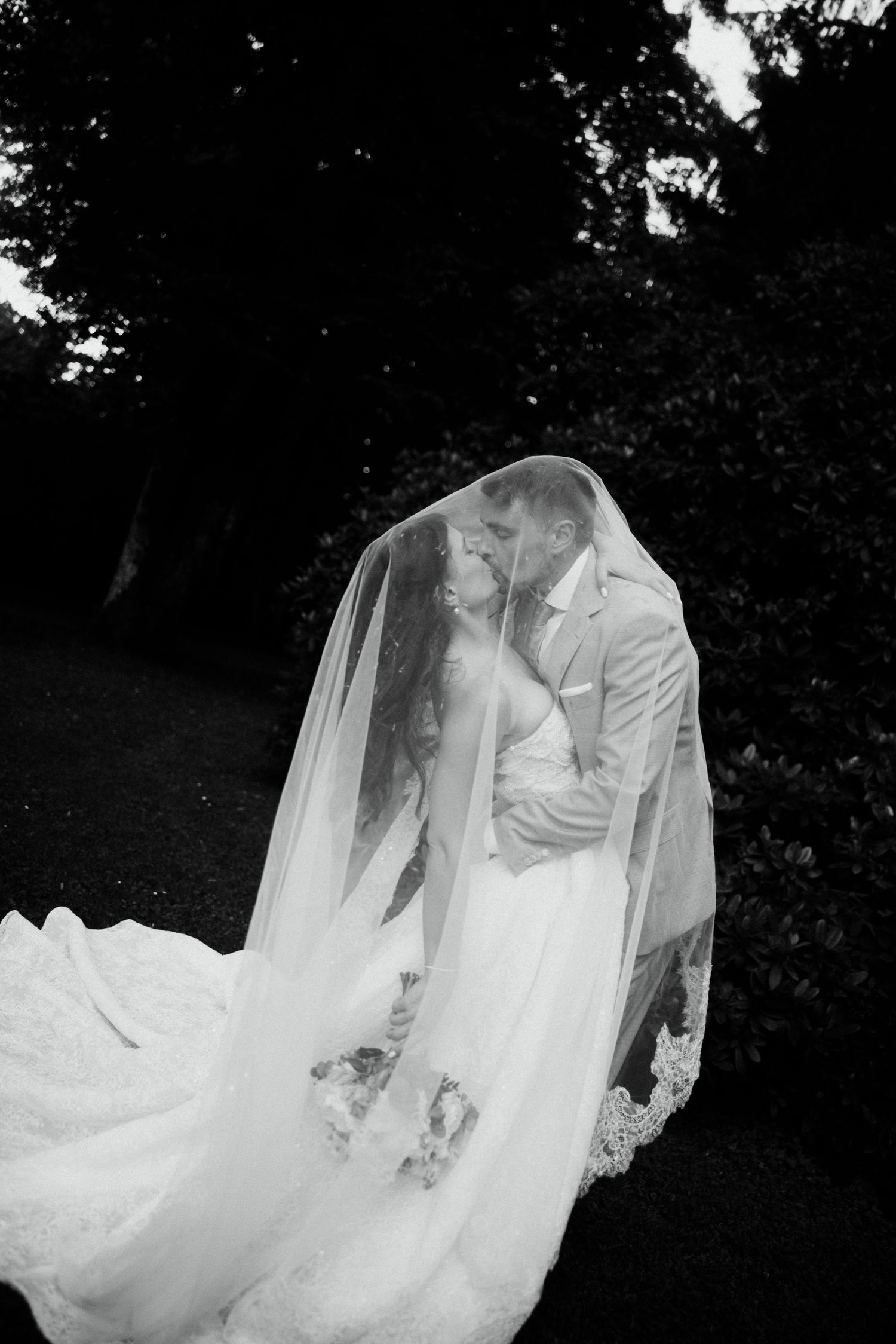 Black and white photo of bride and groom kissing under a flowing veil outdoors.