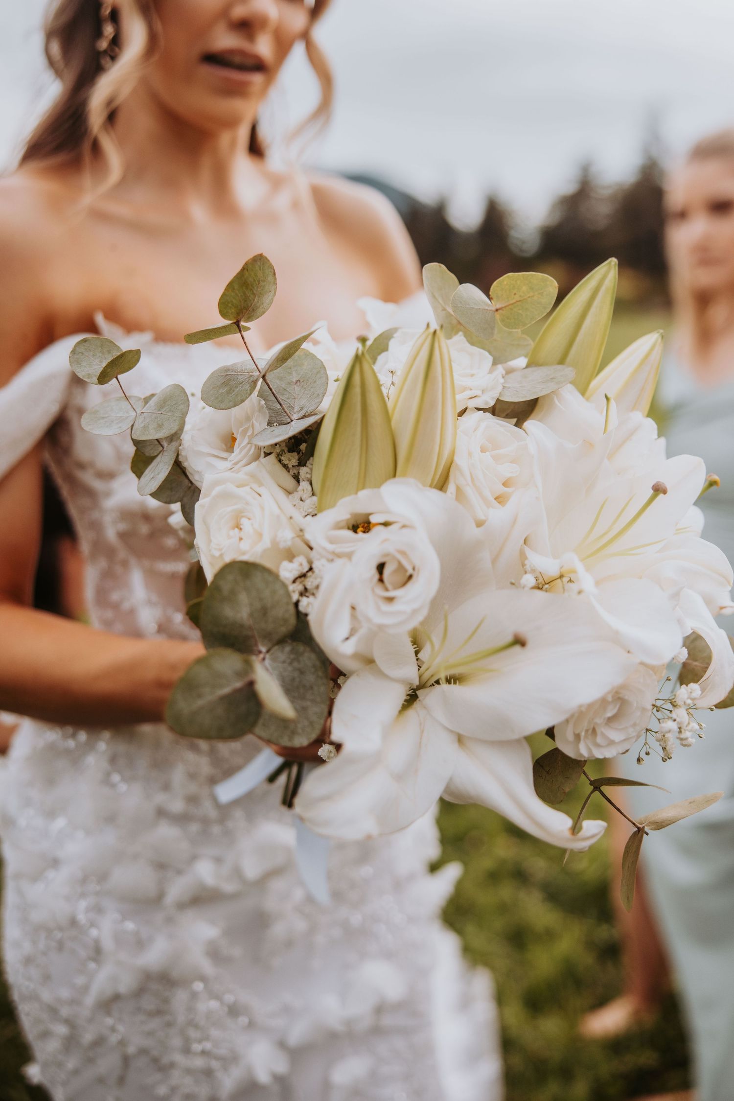 Close-up of bridal bouquet featuring white lilies, roses, and eucalyptus held by bride in lace gown.