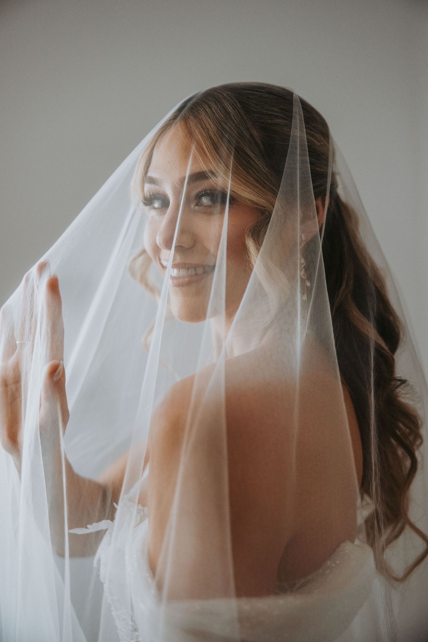 Bride smiling under a sheer white wedding veil, holding it up with her hand in a soft portrait.