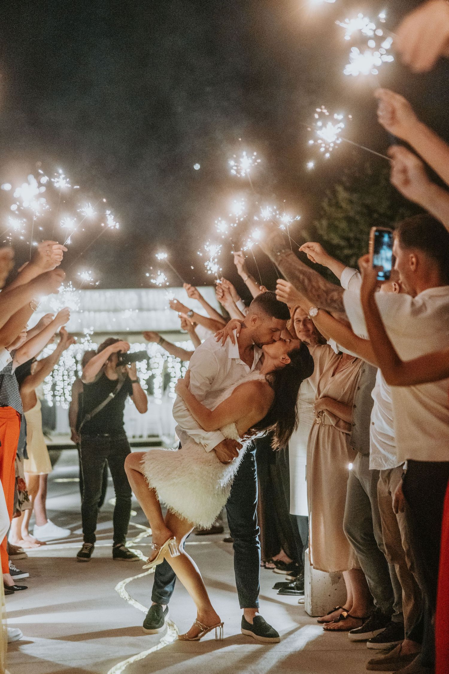 Newlyweds share passionate kiss during sparkler exit surrounded by cheering wedding guests at night.