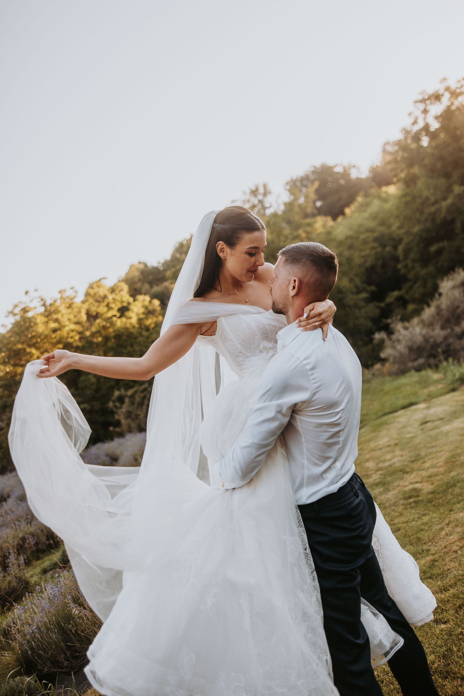 Groom lifting and kissing his bride in a flowing white gown and veil outdoors at sunset.