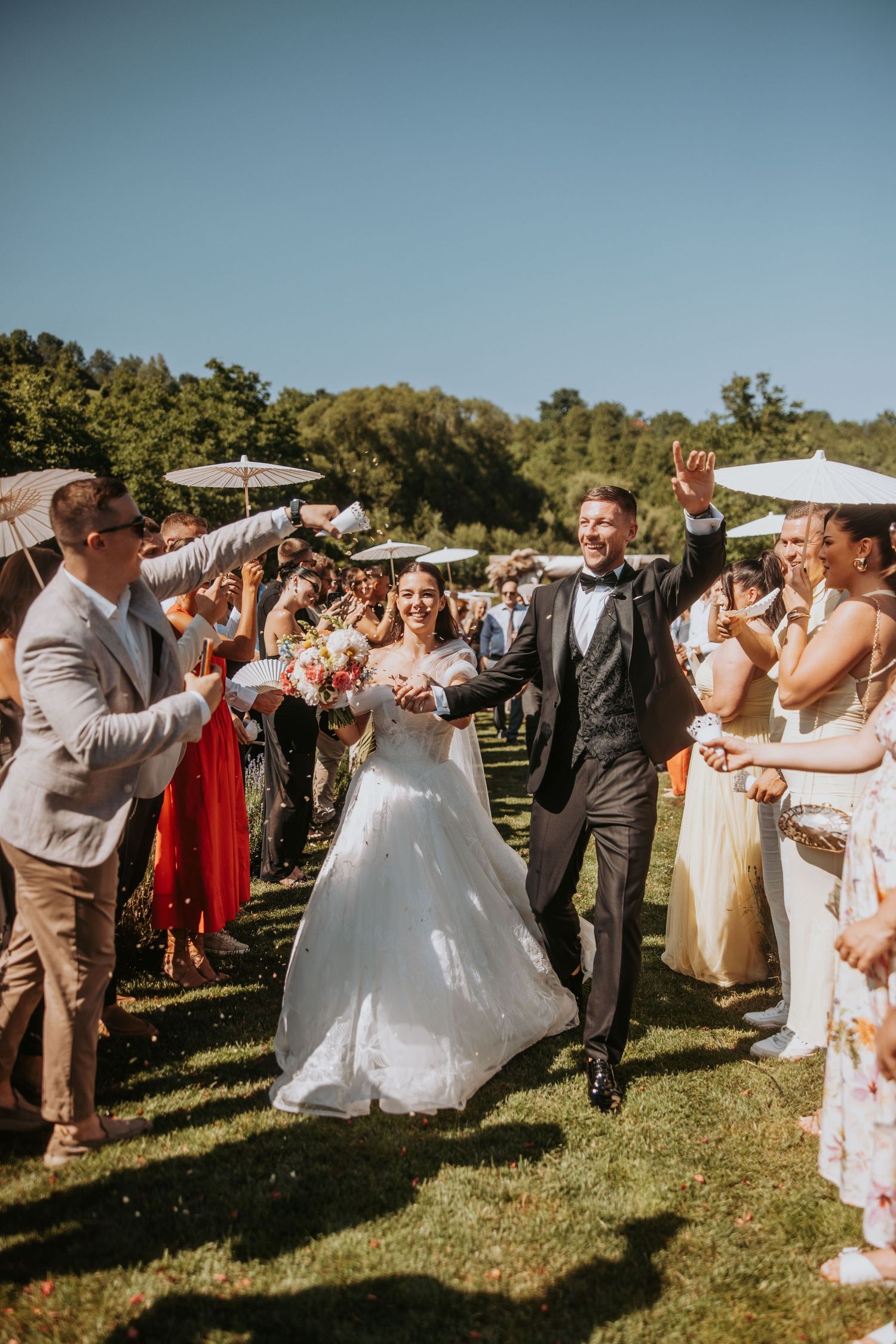 Joyful bride and groom walk through cheering guests holding parasols at an outdoor wedding ceremony.