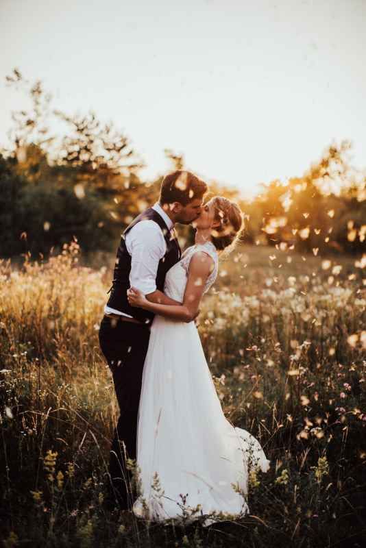 Newlyweds sharing a golden sunset kiss in a wildflower meadow with petals floating around them.