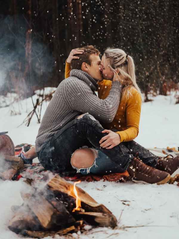 Couple kissing by a campfire in snowy winter woods, wearing cozy sweaters during snowfall.