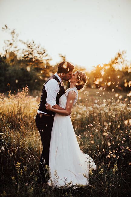 Bride and groom share a sunset kiss in a wildflower meadow with golden bokeh light.