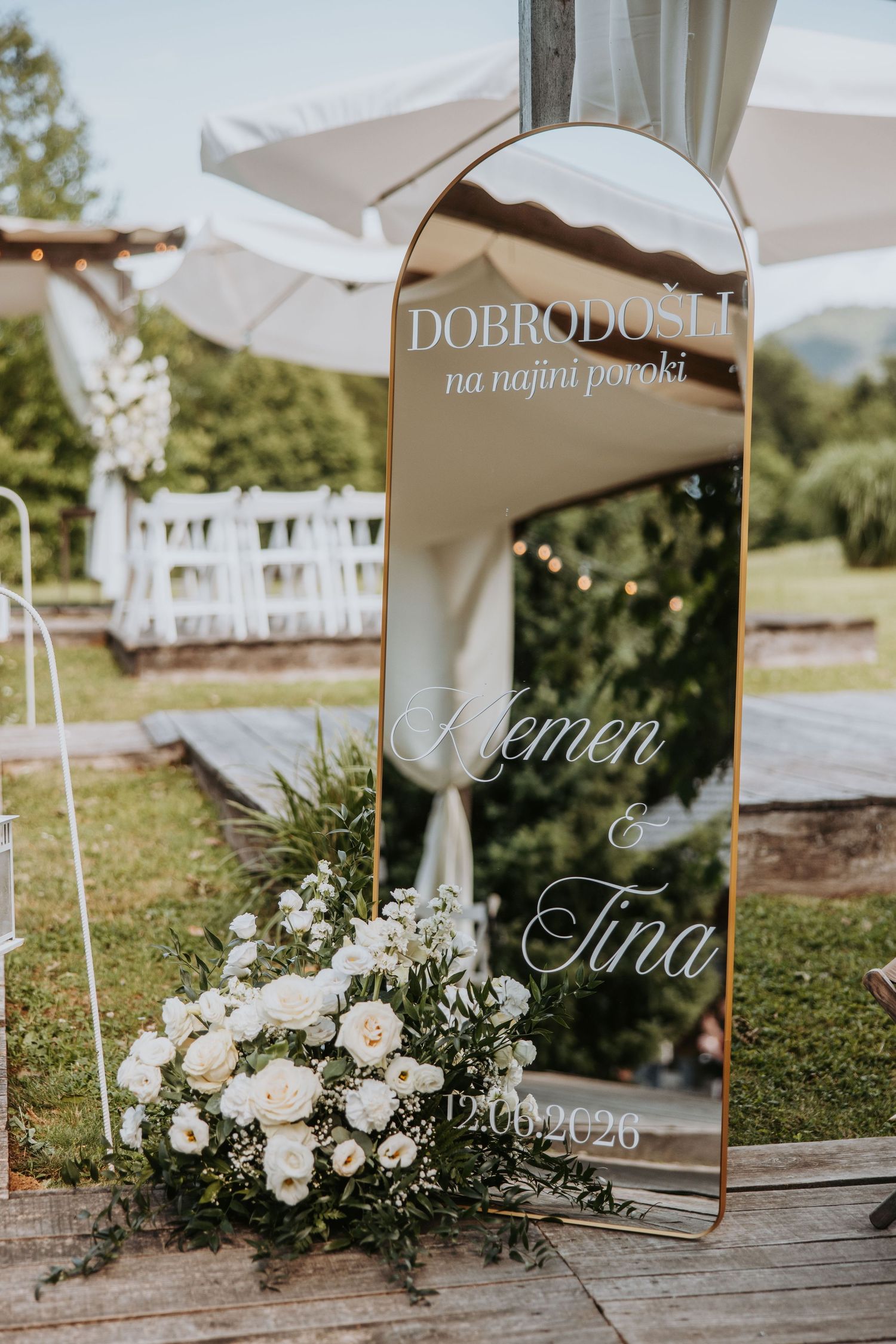 Elegant wedding welcome mirror sign for Klemen and Tina adorned with white floral arrangements outdoors.