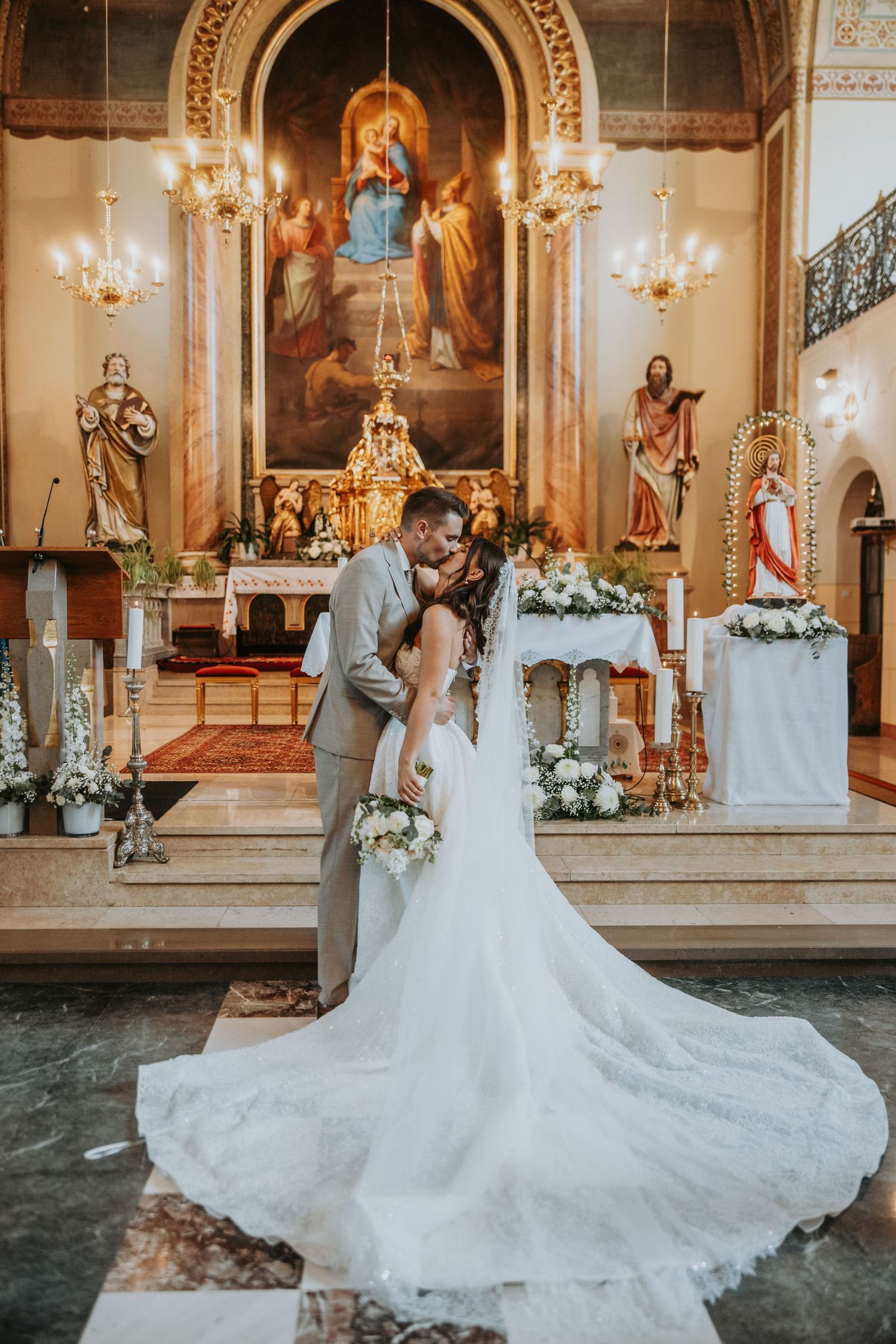 Bride and groom sharing first kiss at decorated church altar with religious paintings backdrop.