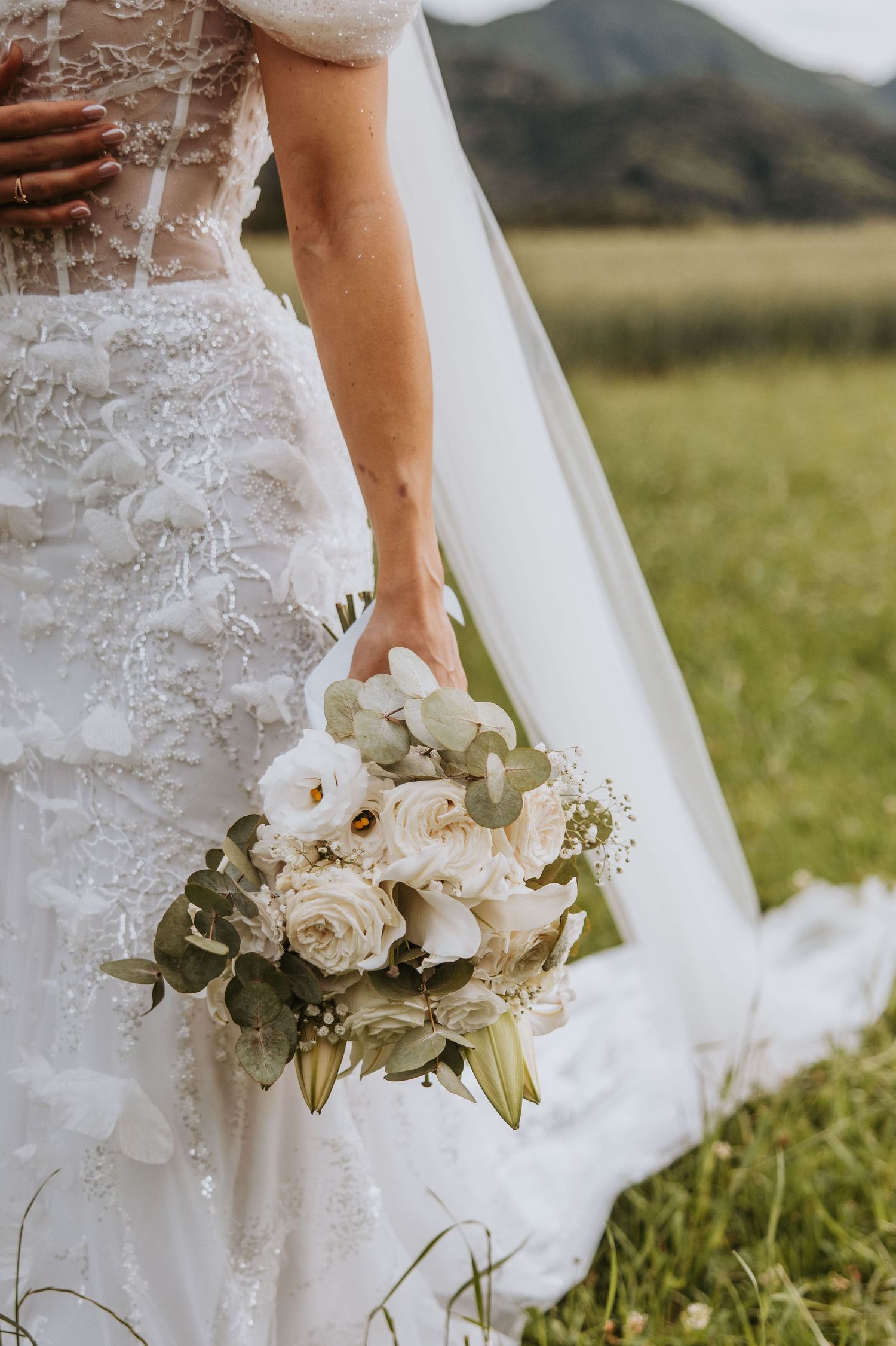 Bride holding elegant white rose and eucalyptus bouquet in lace wedding dress with veil in green meadow.