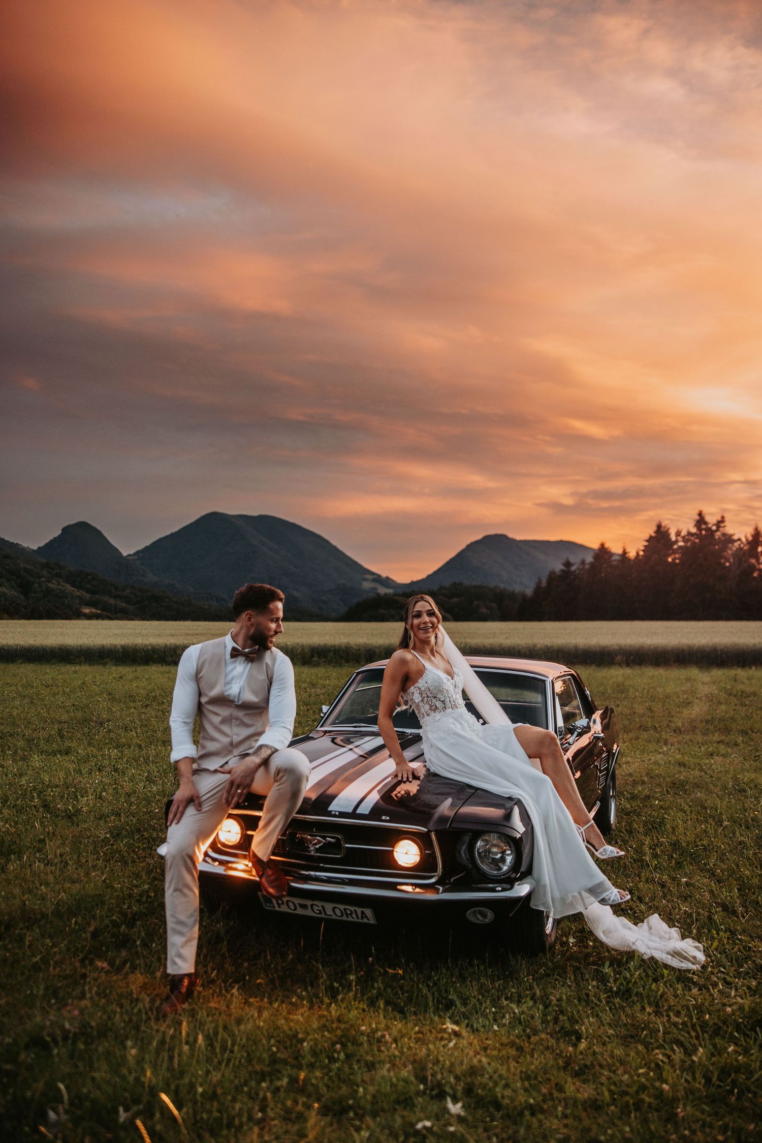 Bride and groom sit on vintage Mustang hood in mountain meadow under breathtaking golden sunset sky.