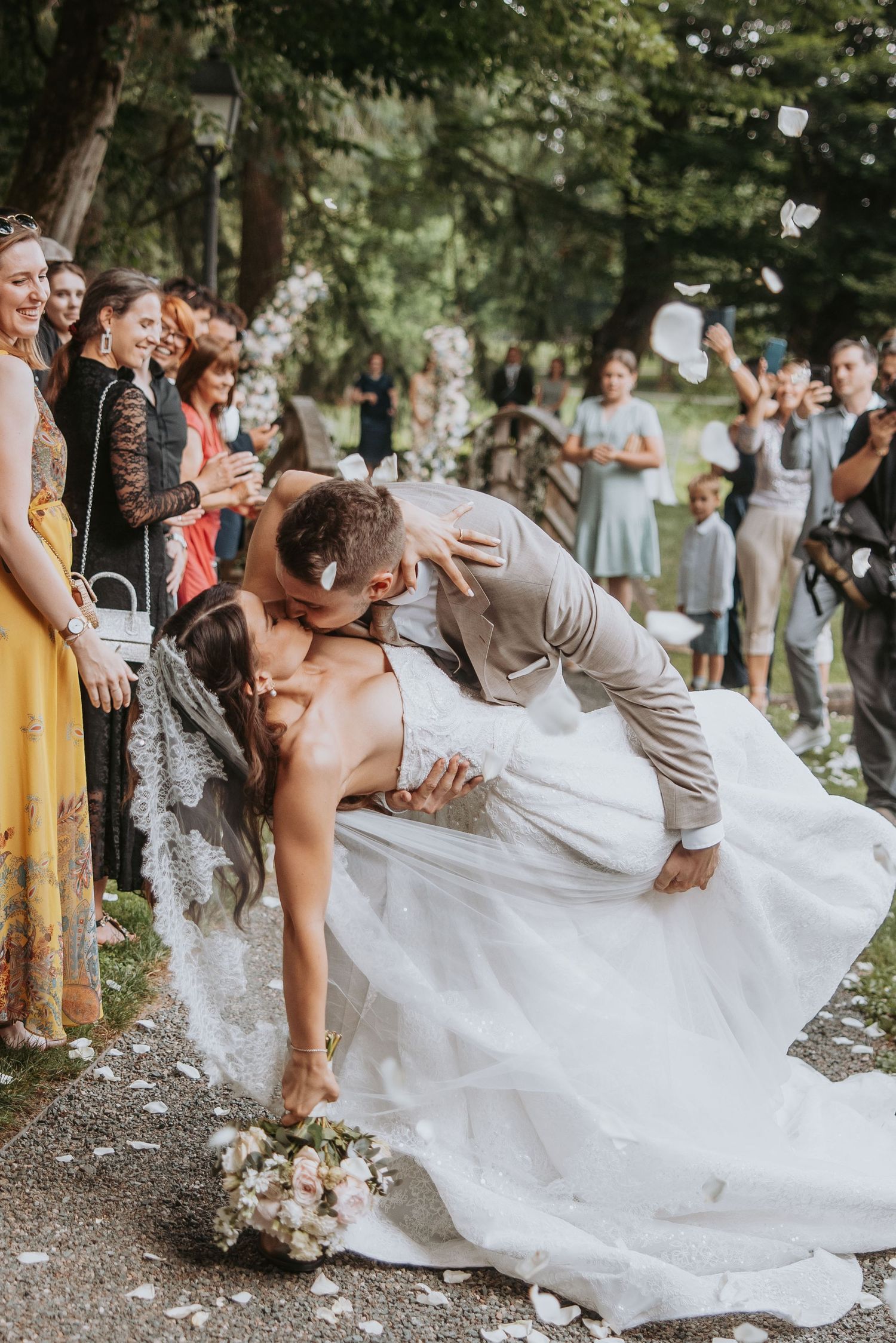 Groom dips bride for passionate kiss while wedding guests celebrate with confetti outdoors.