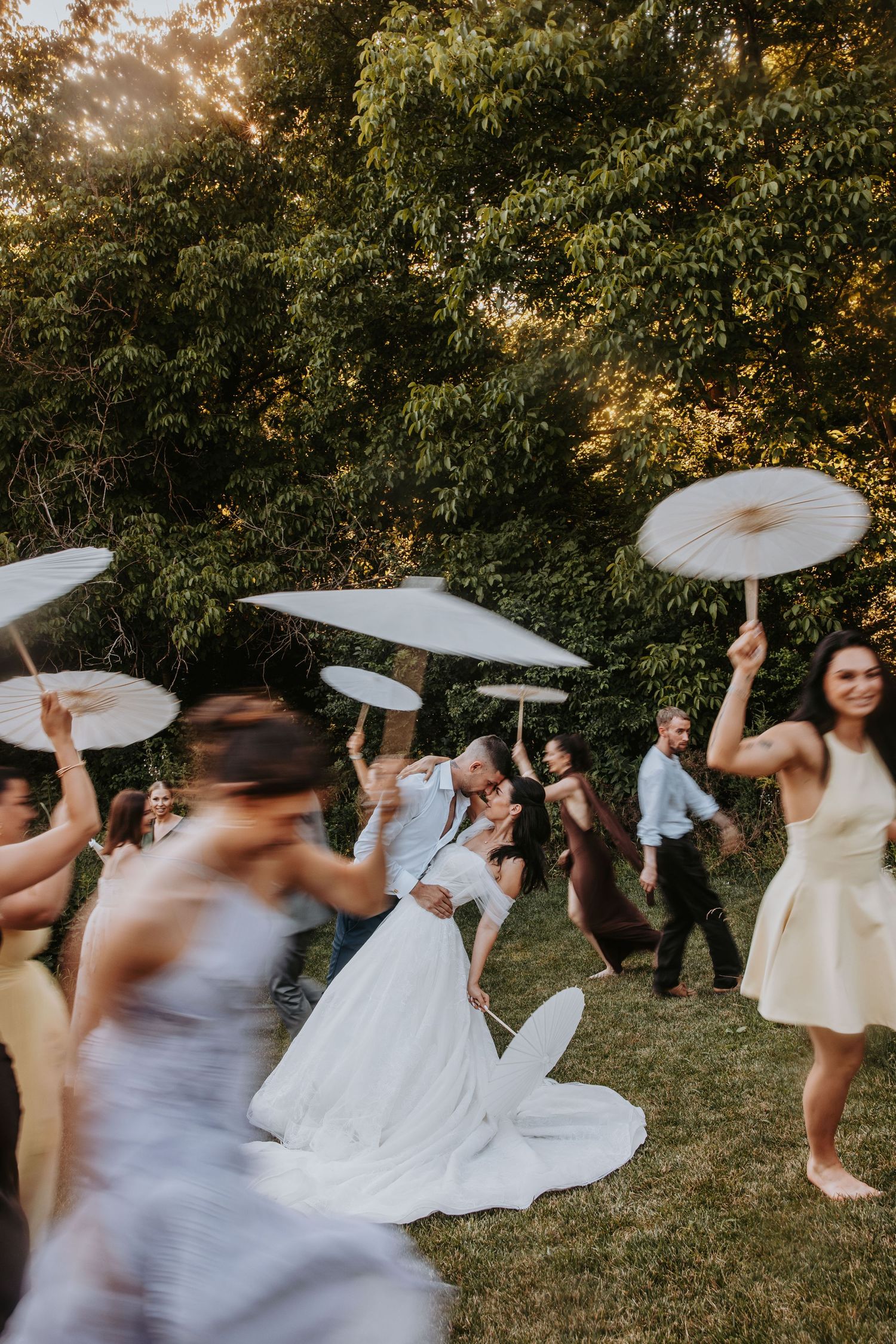 Bride and groom share a dip kiss surrounded by dancing guests waving parasols at an outdoor reception.